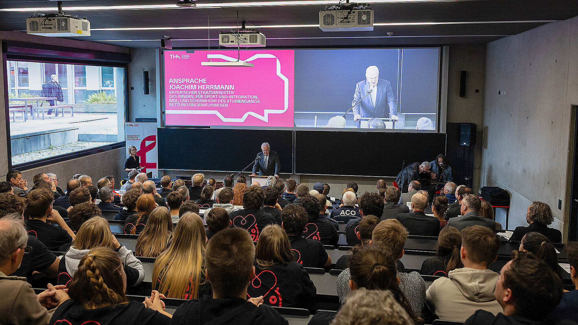 Ein Bild von einer Veranstaltung im Hörsaal, bei der Innenminister Joachim Herrmann am Rednerpult spricht. Auf der großen Leinwand im Hintergrund ist eine Präsentation mit dem Titel "Ansprache Joachim Herrmann" zu sehen. Der Saal ist voll mit Zuhörern, die aufmerksam dem Vortrag folgen.