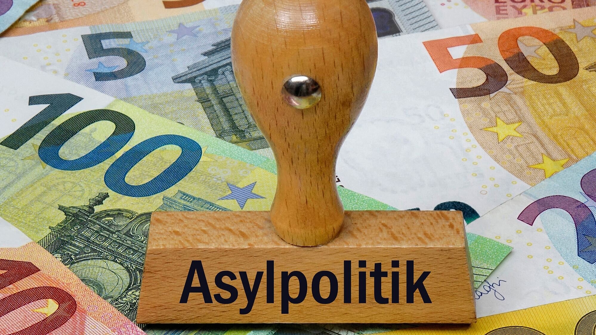 Auf dem Bild ist ein Stempel mit der Aufschrift "Asylpolitik" zu sehen, darunter liegen mehrere Geldscheine.