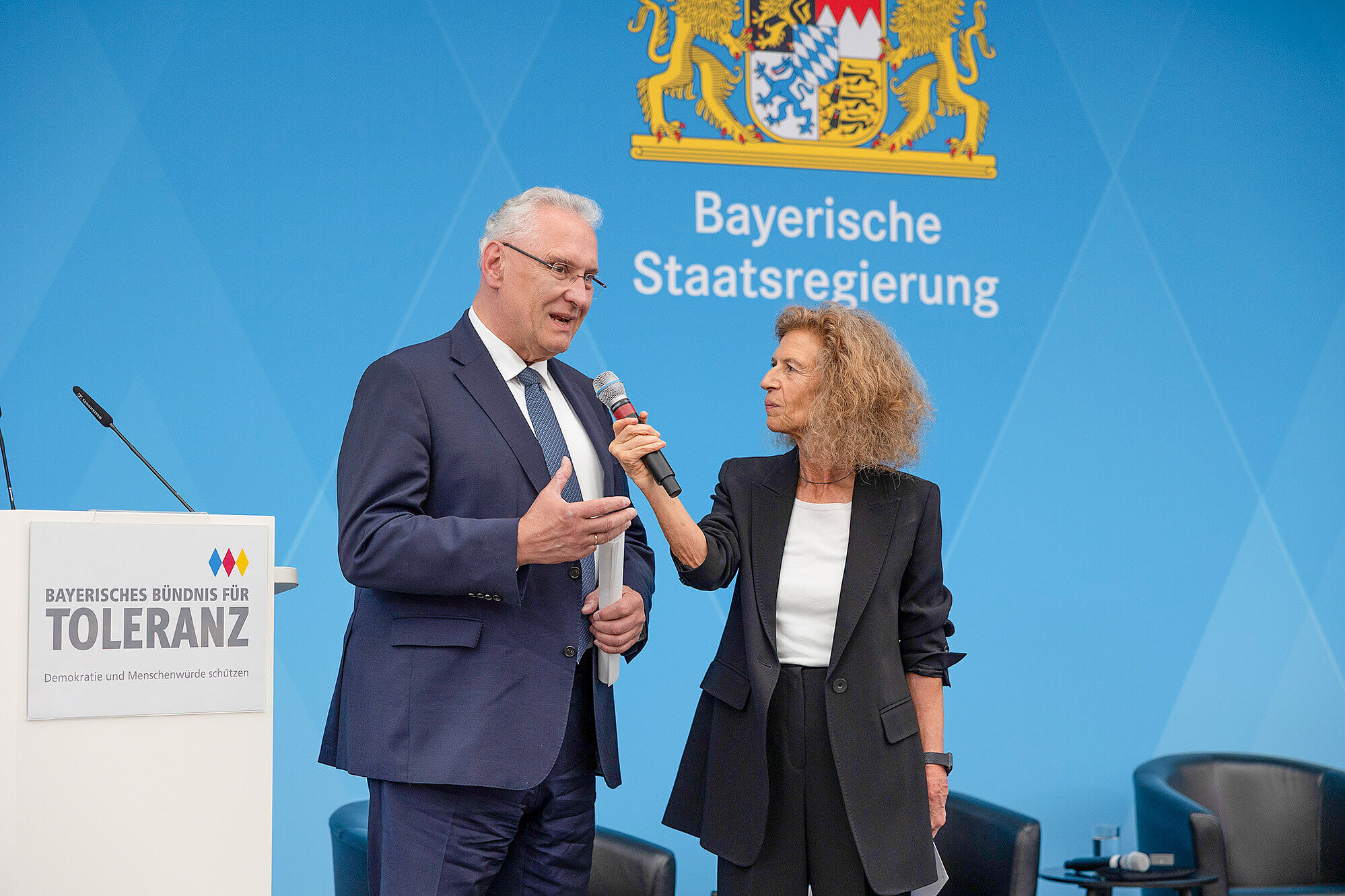 links Innenminister Joachim Herrmann rechts Moderatorin hält ein Mikrofon Richtung Joachim Herrmann der zum Publikum spricht im Hintergrund ist das Wappen der Bayerischen Staatsregierung auf einer blauen Wand