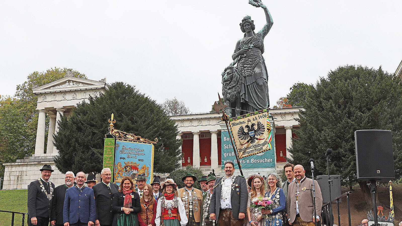 Auf dem Bild sind Menschen bei der Feier zur Siegerehrung der Landeschützen vor der Bavaria zu sehen. Die Statue zeigt eine Frau mit erhobener Hand, die ein Schwert hält, und steht auf einem Sockel. Die Menschen tragen festliche Kleidung, einige sind in traditionellen Trachten. Zwei Banner sind sichtbar, eines mit einem Wappen und Text, das andere mit einem schwarzen Adler. Im Hintergrund sind Säulen und ein Gebäude mit Säulen zu erkennen. Die Szene wirkt feierlich, vermutlich bei einer Ehrung oder einem Fest.