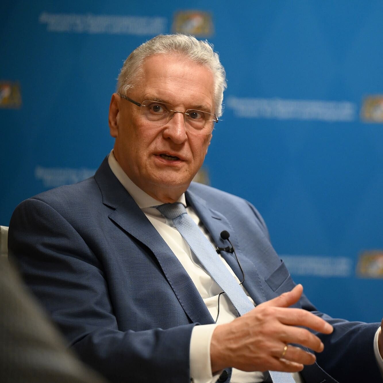 Auf dem Bild ist Innenminister Joachim Herrmann zu sehen, wie er in einer Gesprächsrunde diskutiert. Hinter ihm ist eine Zipperwand des Innenministeriums aufgebaut.