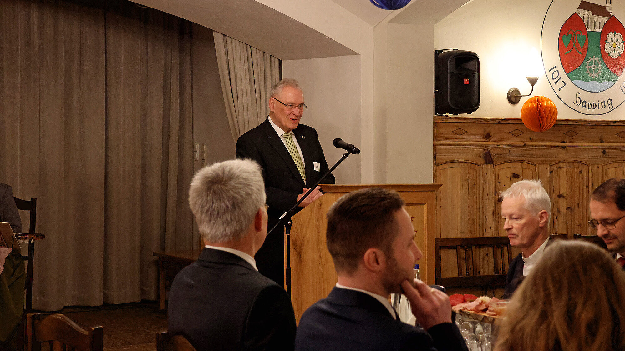 Innenminister Joachim Herrmann im Anzug steht an einem hölzernen Rednerpult vor einem Mikrofon und hält eine Rede in einem gemütlichen Gastraum; mehrere Personen sitzen an gedeckten Tischen und hören zu. An der Wand ist ein Wappen, von der Decke hängen Papierdekorationen.