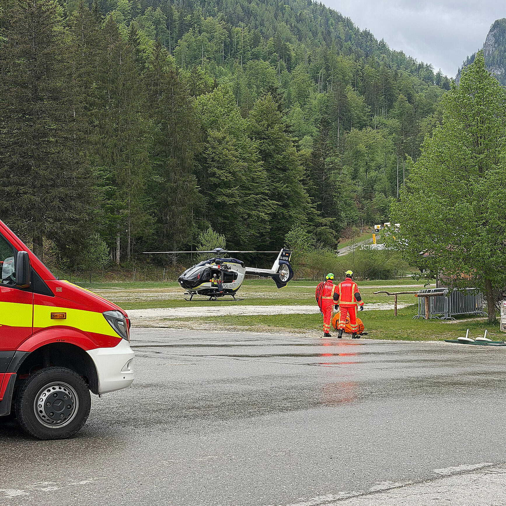 Rettungshubschrauber auf einer Wiese vor bewaldeten Bergen; zwei Rettungsdienst Personen in orangefarbener Schutzkleidung, ein Rettungswagen am Straßenrand, nasser Asphalt, bewölkter Himmel.