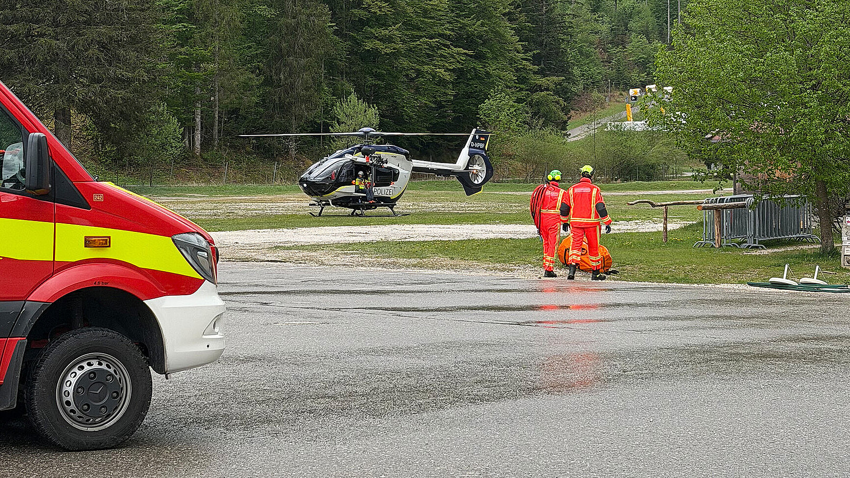 Rettungshubschrauber auf einer Wiese vor bewaldeten Bergen; zwei Rettungsdienst Personen in orangefarbener Schutzkleidung, ein Rettungswagen am Straßenrand, nasser Asphalt, bewölkter Himmel.