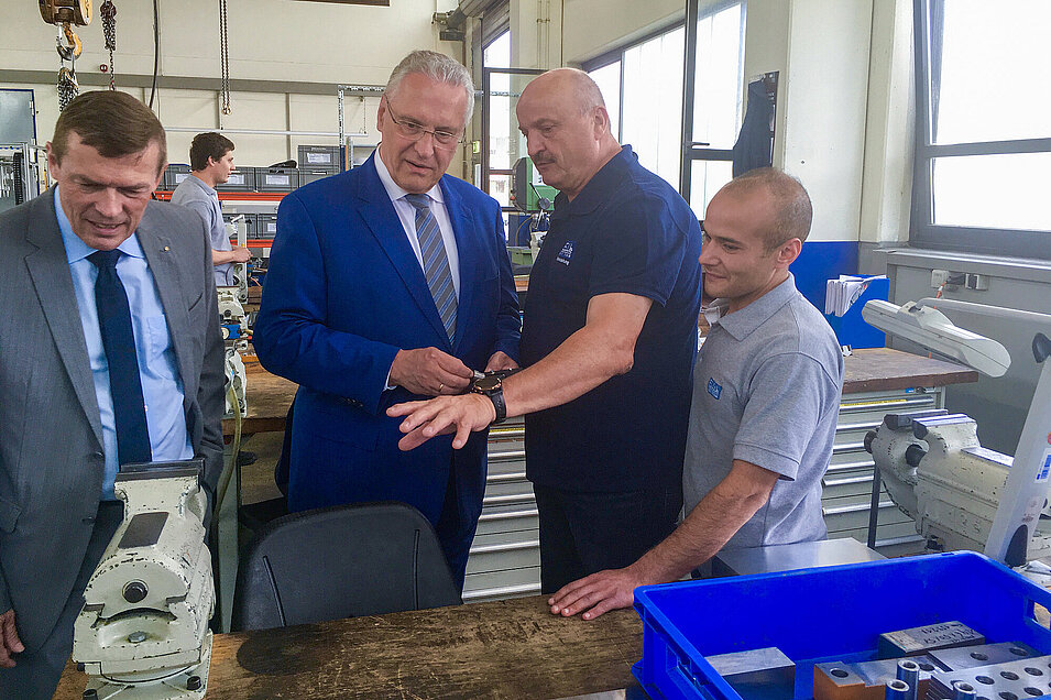 Innenminister Joachim Herrmann im Gespräch mit Arbeitern an der Werkbank, davon ein Arbeiter mit Migrationshintergrund