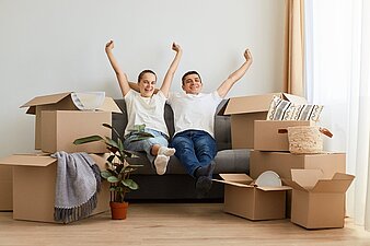 Inmitten von Umzugskartons sitzen ein Mann und eine Frau auf einem Sofa und reißen erfreut die Hände nach oben