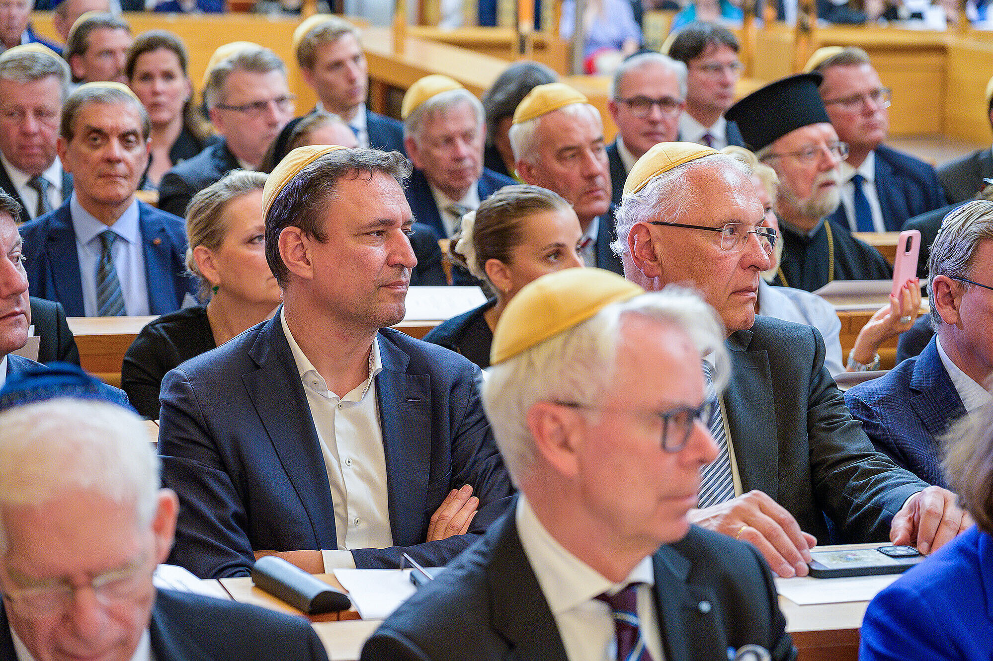 Publikum mit Blick zur Bühne in Reihe zwei sitzen Justizminister Georg Eisenreich und Innenminister Joachim Herrmann, sie blicken zur Bühne und tragen beide eine gelbe Kippa.