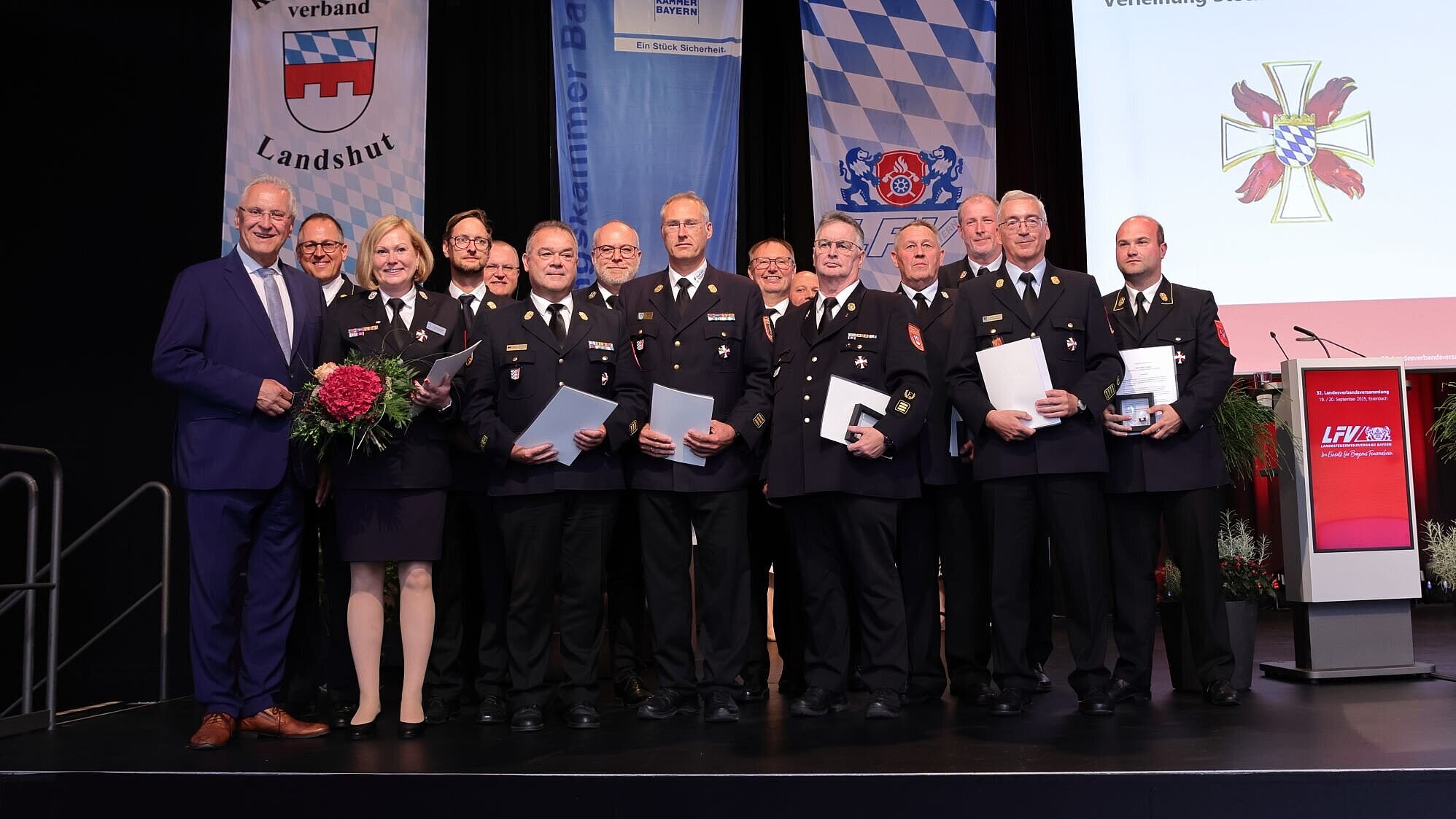 Auf einer Bühne stehen Innenminister Joachim Herrmann, Verbandsvertreter des LFV Bayern sowie die Feuerwehr-Angehörigen, die mit dem Steckkreuz ausgezeichnet worden sind. Alle lächeln zur Kamera.