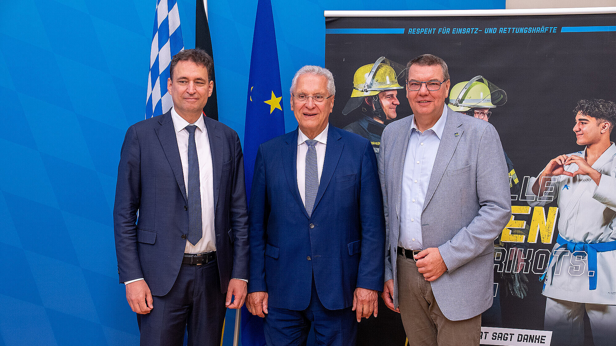 Justizminister Georg Eisenreich, Innenminister Joachim Herrmann und den Vorsitzenden der Bayerischen Polizeistiftung Thomas Lintl stehen vor einer brauen Pressewand, Fahnen, sowie einen Roll-up zur Kampagne Eure Fan.