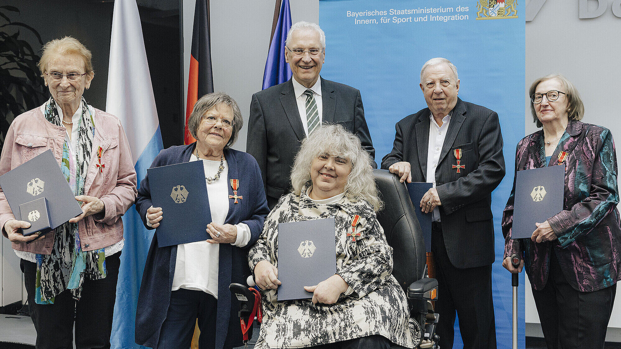 Zu sehen sind Innenminister Joachim Herrmann und die Beliehenen mit ihren Orden und Urkunden, hinter ihnen Fahnen und ein Roll-Up.