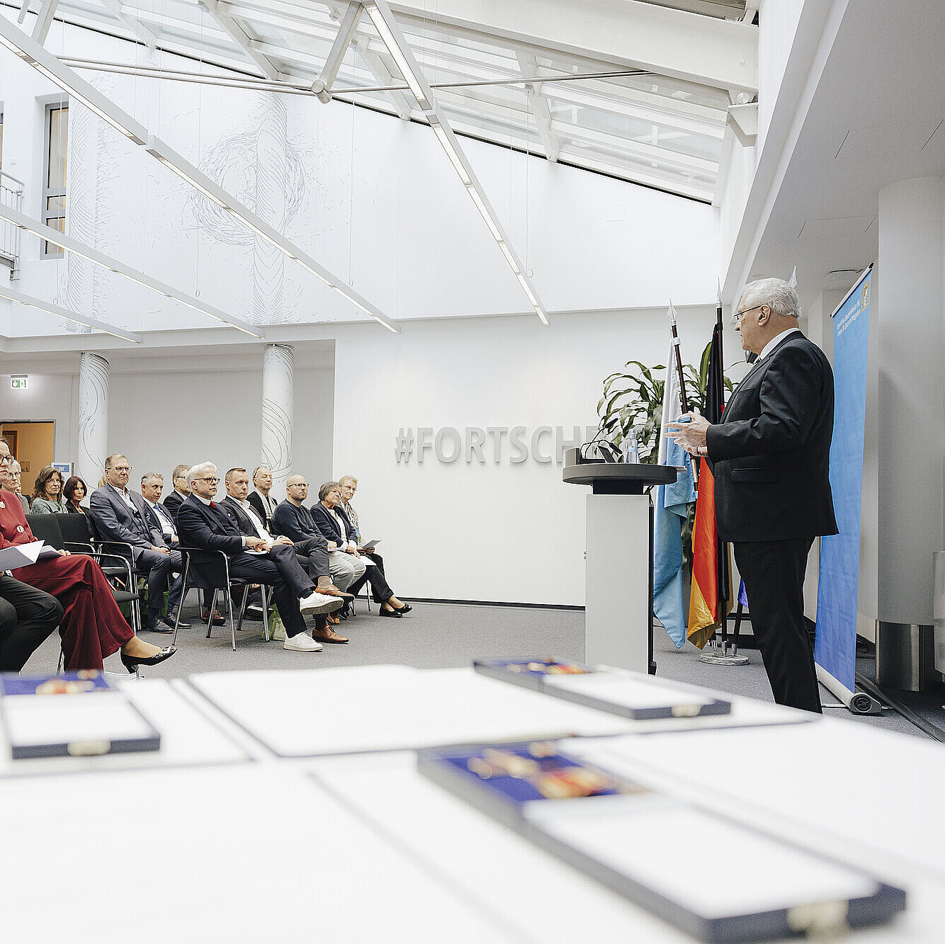 Auf dem Bild ist Innenminister Joachim Herrmann am Rednerpult während seiner Rede zu sehen, das Publikum hört ihm zu. Im Vordergrund sieht man noch die Verdienstkreuze.