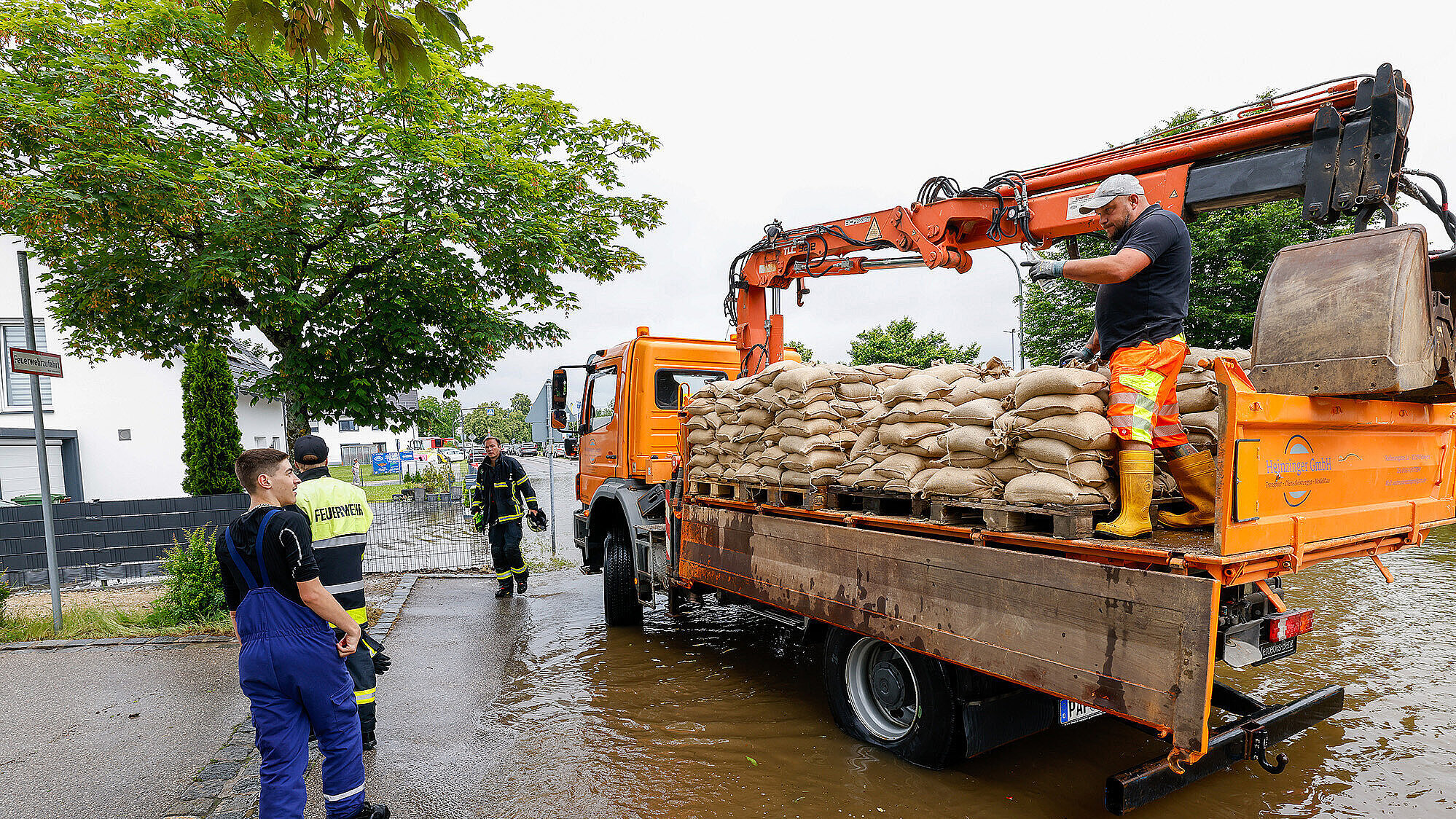 Das Bild zeigt einen Einsatz bei starkem Regen, bei dem Feuerwehrkräfte und Helfer mit einem orangefarbenen Einsatzfahrzeug auf einer überschwemmten Straße arbeiten. Ein Mann steht auf dem Fahrzeug, das mit Sandsäcken beladen ist, und überprüft die Ausrüstung. Zwei weitere Helfer in Feuerwehruniformen stehen am Straßenrand, während Wasser die Straße überschwemmt. Im Hintergrund sind Bäume, Gebäude und Einsatzfahrzeuge zu sehen.