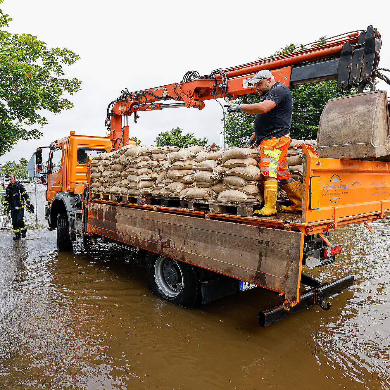 Das Bild zeigt einen Einsatz bei starkem Regen, bei dem Feuerwehrkräfte und Helfer mit einem orangefarbenen Einsatzfahrzeug auf einer überschwemmten Straße arbeiten. Ein Mann steht auf dem Fahrzeug, das mit Sandsäcken beladen ist, und überprüft die Ausrüstung. Zwei weitere Helfer in Feuerwehruniformen stehen am Straßenrand, während Wasser die Straße überschwemmt. Im Hintergrund sind Bäume, Gebäude und Einsatzfahrzeuge zu sehen.