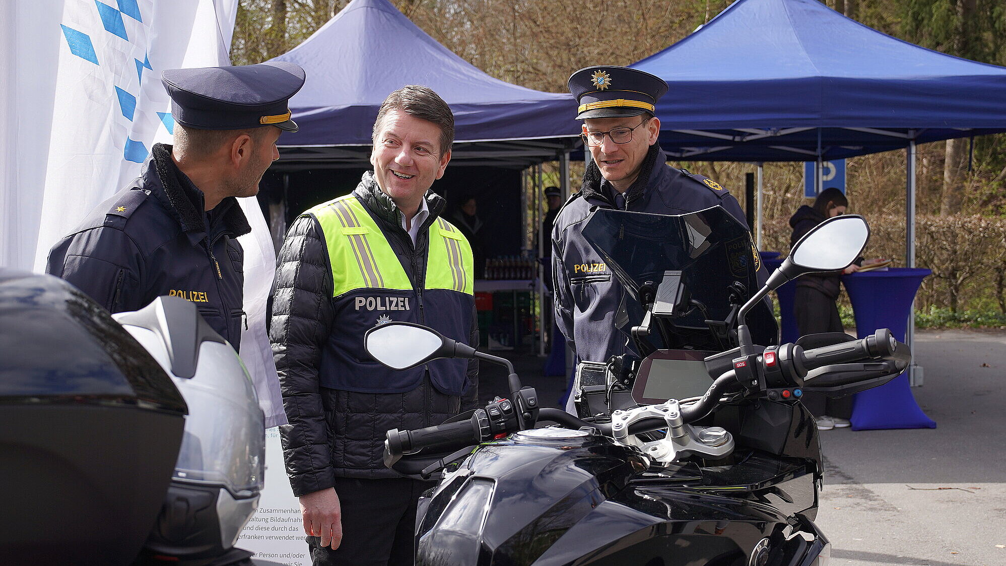 Drei Männer im Gespräch, zwei davon in Polizeiuniform, im Vordergrund ein Motorrad