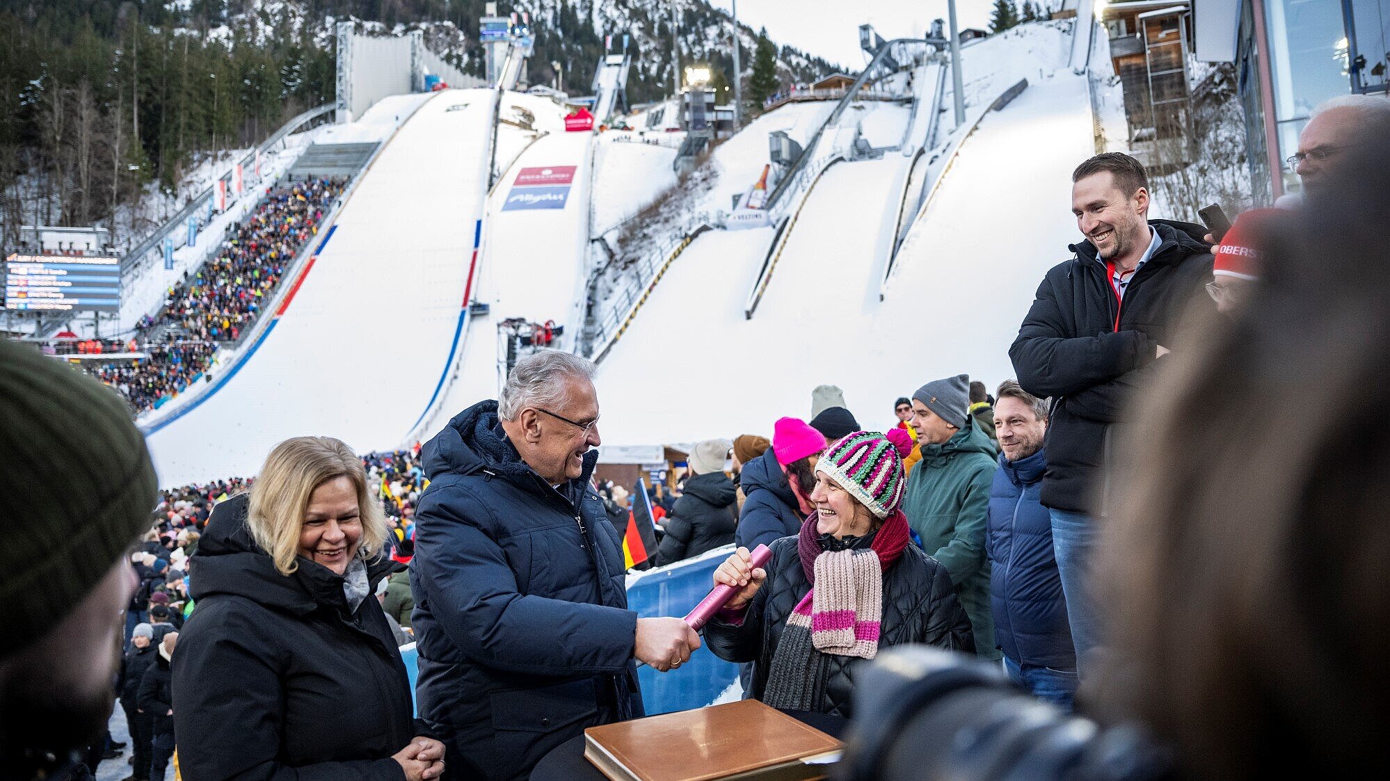 Herrmann übergibt neben Bundesinnenministerin Faeser Stab an die baden-württembergische Kultus-, Jugend- und Sportministerin Theresa Schopper, im Hintergrund Skisprungschanze von Oberstdorf