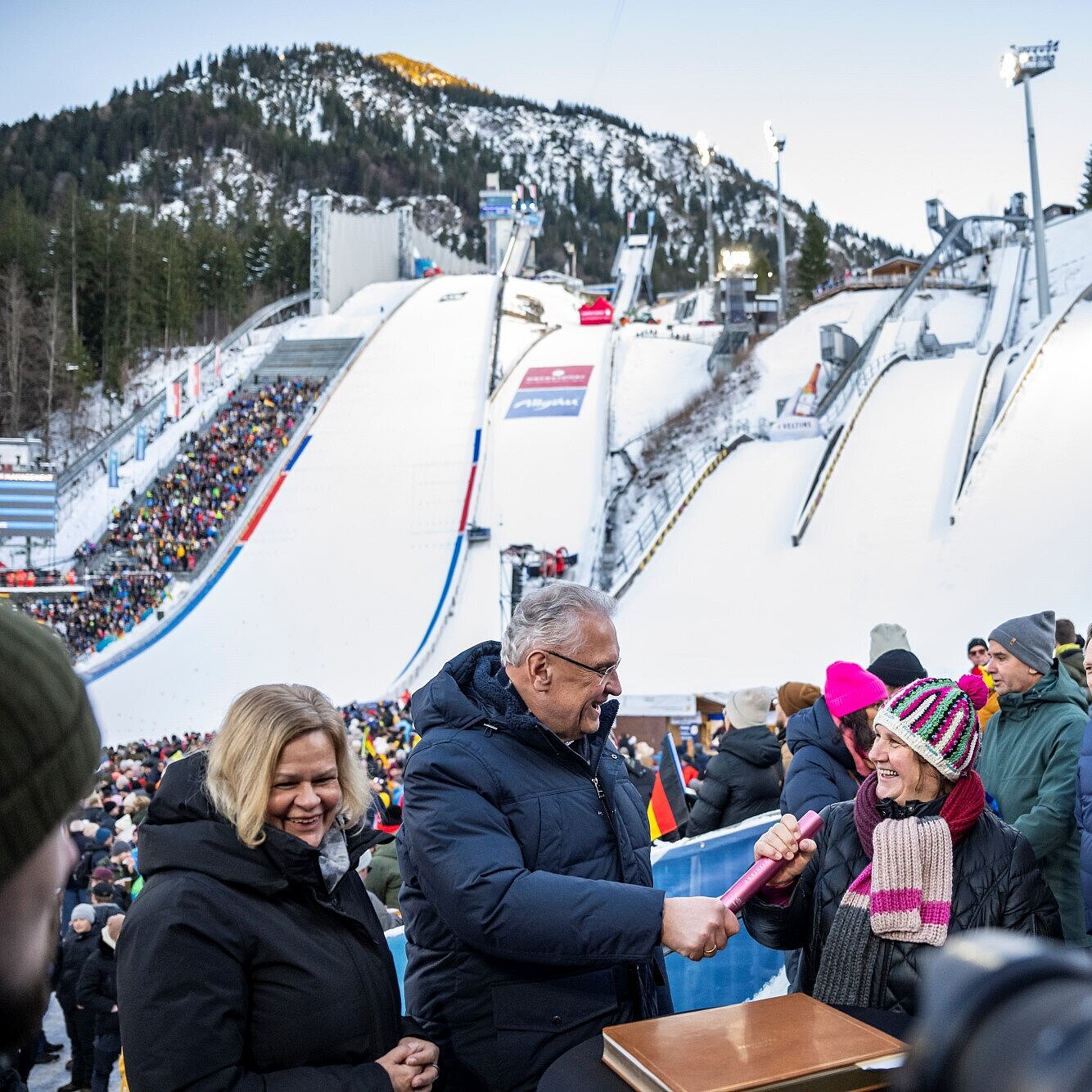 Herrmann übergibt neben Bundesinnenministerin Faeser Stab an die baden-württembergische Kultus-, Jugend- und Sportministerin Theresa Schopper, im Hintergrund Skisprungschanze von Oberstdorf