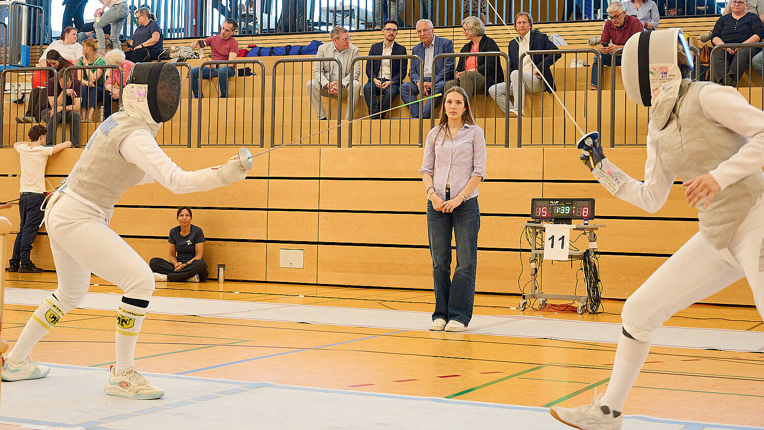 Zwei Fechter in weißer Schutzkleidung stehen im Ausfallschritt auf der Planche einer Sporthalle, im Hintergrund sitzen Zuschauer auf Holzbänken und weitere Teilnehmer betreten die Bahnen.