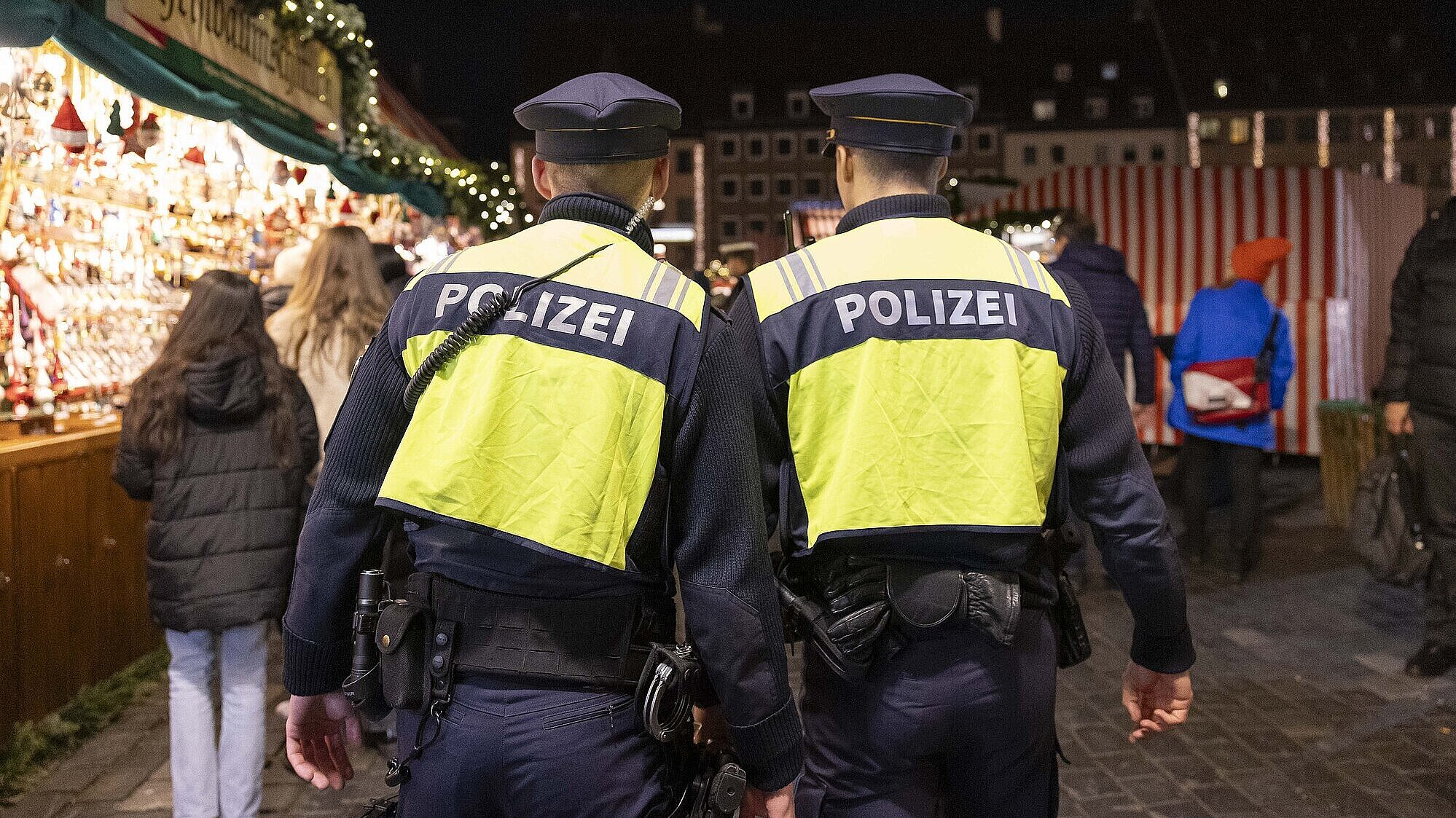 Zwei Polizisten in gelben Warnwesten mit der Aufschrift „POLIZEI“ gehen bei Nacht über einen belebten Christkindlesmarkt in Nürnberg, im Hintergrund sind Marktstände mit Weihnachtsdekoration und mehrere Besucher zu sehen.