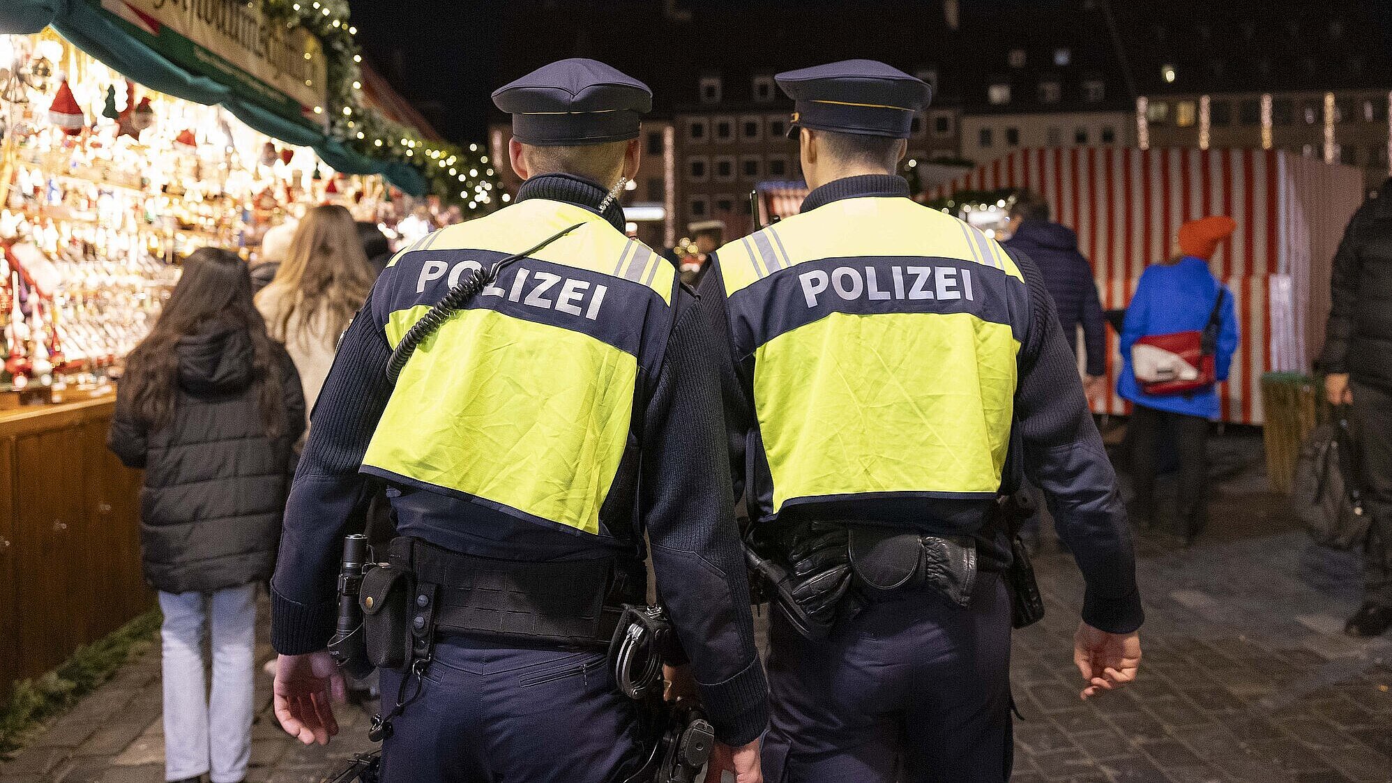 Zwei Polizisten in gelben Warnwesten mit der Aufschrift „POLIZEI“ gehen bei Nacht über einen belebten Christkindlesmarkt in Nürnberg, im Hintergrund sind Marktstände mit Weihnachtsdekoration und mehrere Besucher zu sehen.