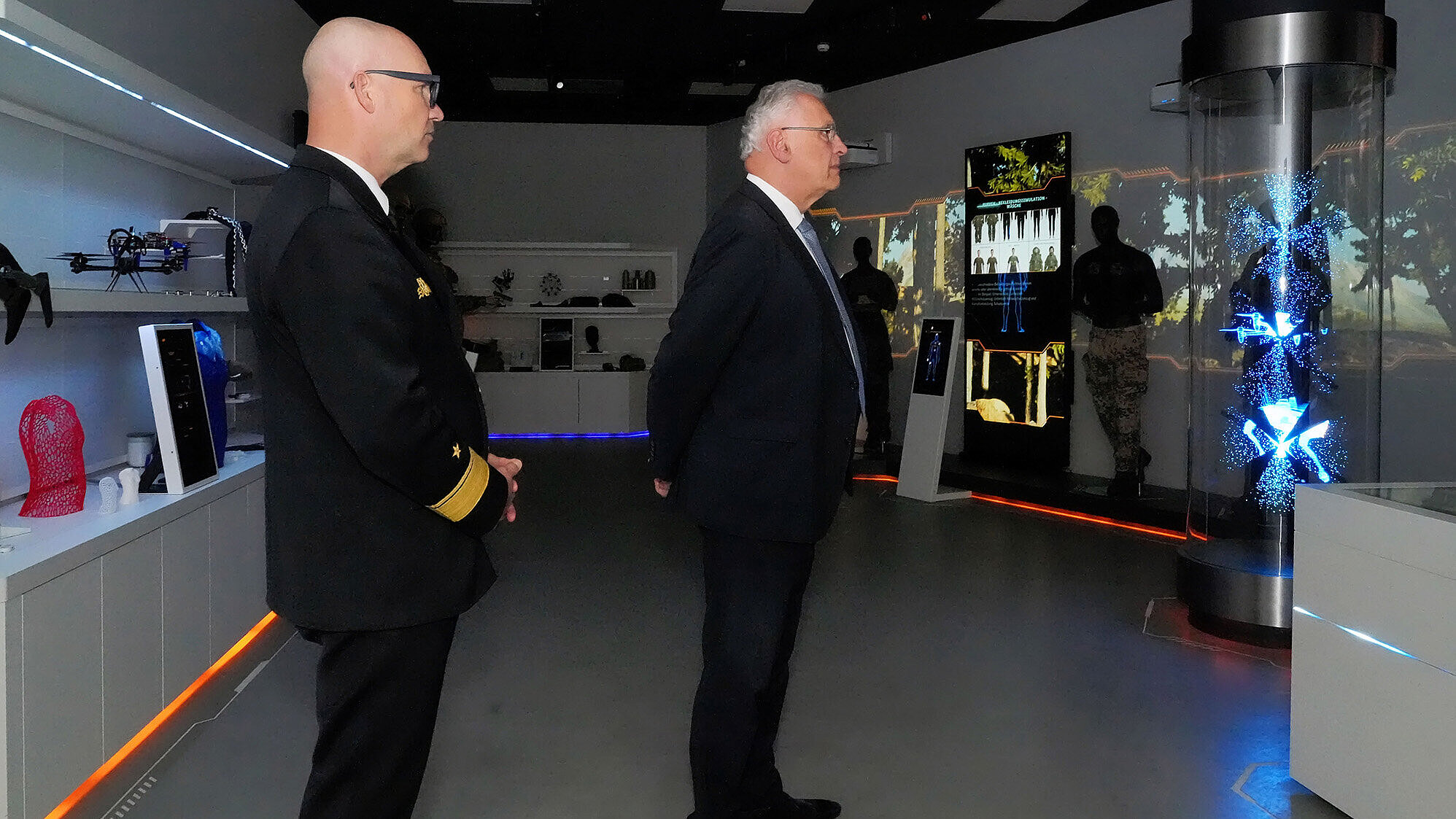 Zwei Männer in formeller Kleidung – einer in dunkelblauer Uniform mit goldenen Ärmelstreifen, der andere im dunklen Anzug – stehen in einem dunklen, modernen Ausstellungsraum mit leuchtenden Displays und einer zylindrischen Installation mit blauen Lichtprojektionen.