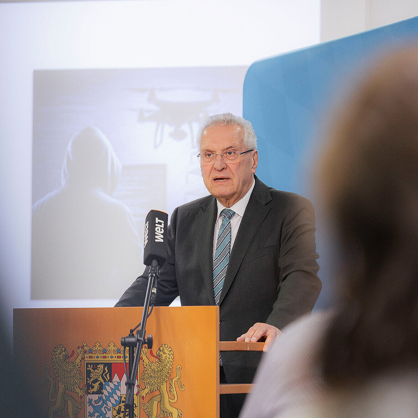 Innenminister Joachim Herrmann in einem dunklen Anzug mit gestreifter Krawatte steht an einem Rednerpult mit dem bayerischen Staatswappen. Vor ihm ist ein Mikrofon mit dem Logo „WELT“ angebracht. Im Hintergrund ist eine Projektion zu sehen, die eine Person mit Kapuzenpullover und eine Drohne zeigt. Im Vordergrund sind unscharf Personen zu erkennen, die dem Vortrag zuhören.