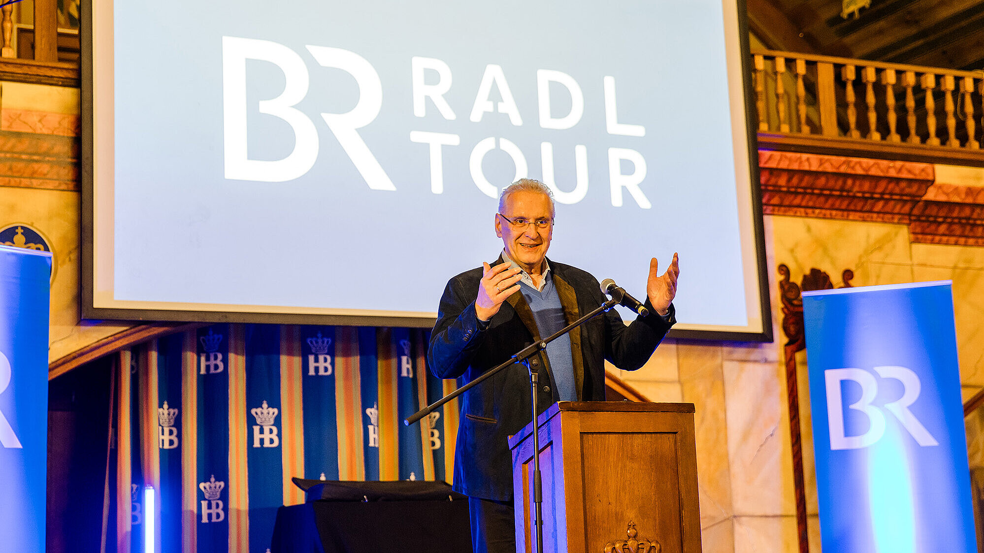 auf dem Bild: Innenminister Herrmann am Rednerpult im Hintergrund ein Bildschirm mit der Aufschrift BR Radl Tour