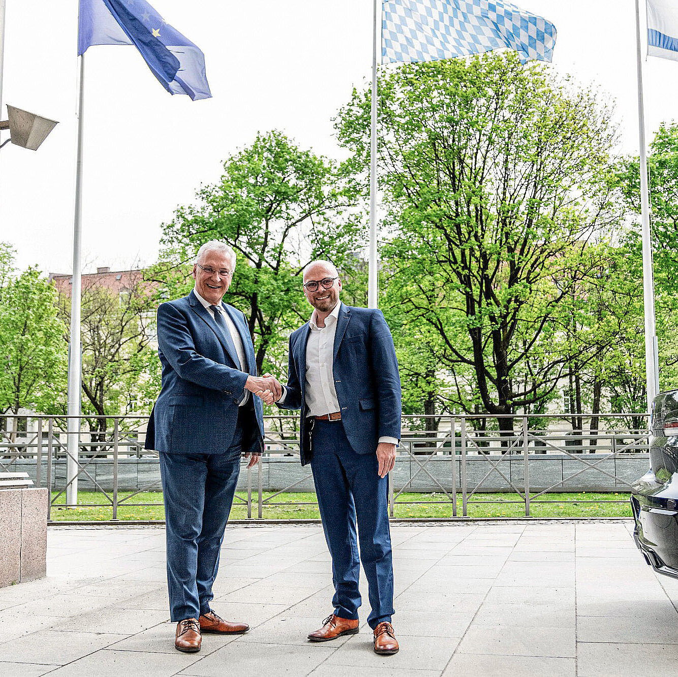 Herrmann und Mehring schütteln sich die Hand vor Fahnenmasten vor Staatskanzlei