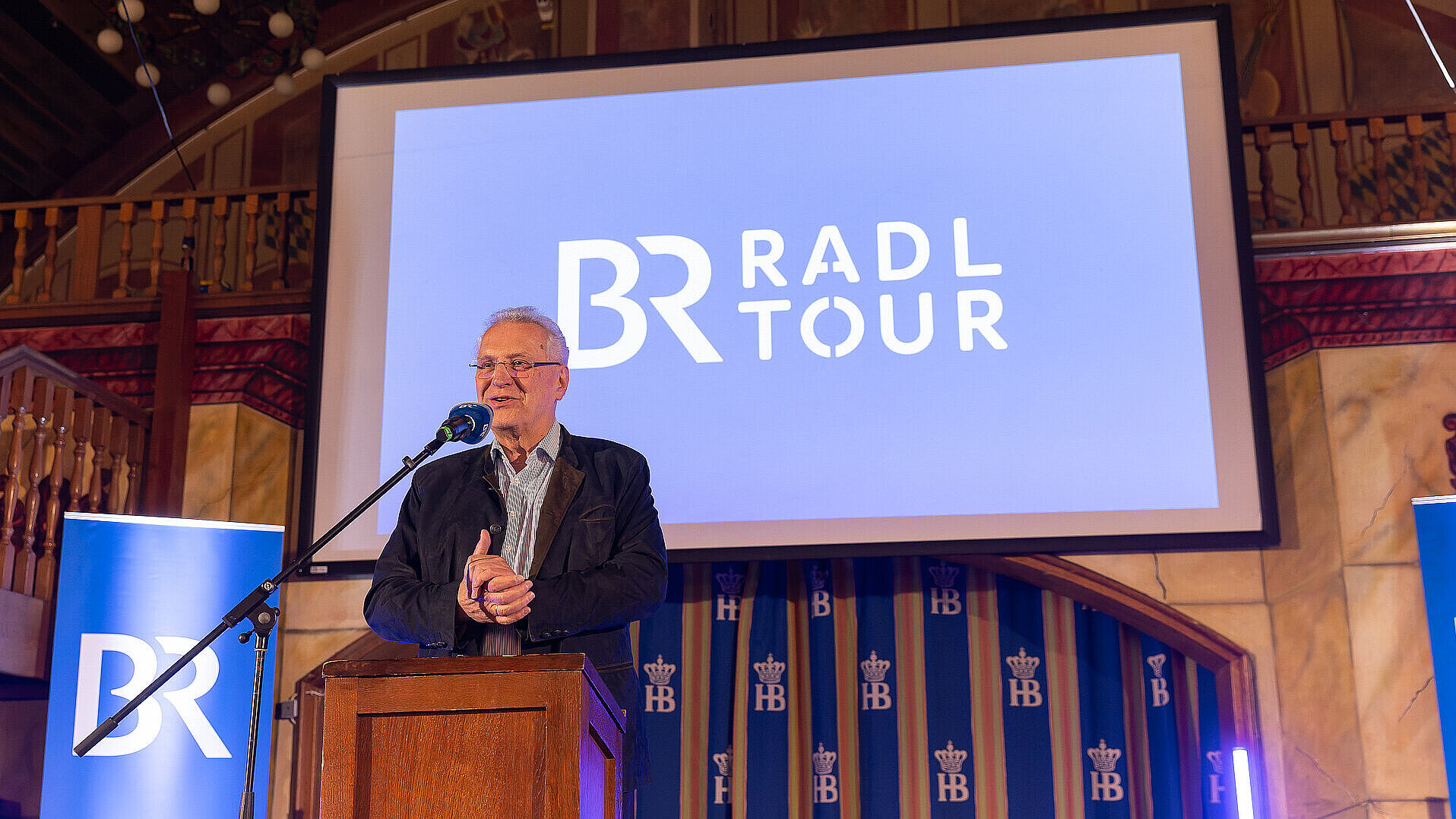 Innenminister Herrmann freundlich lächelnd am Rednerpult, hinter ihm blaue Bildschirmpräsentation zur BR-Radltour, links neben ihm blaues Roll-Up von BR. 