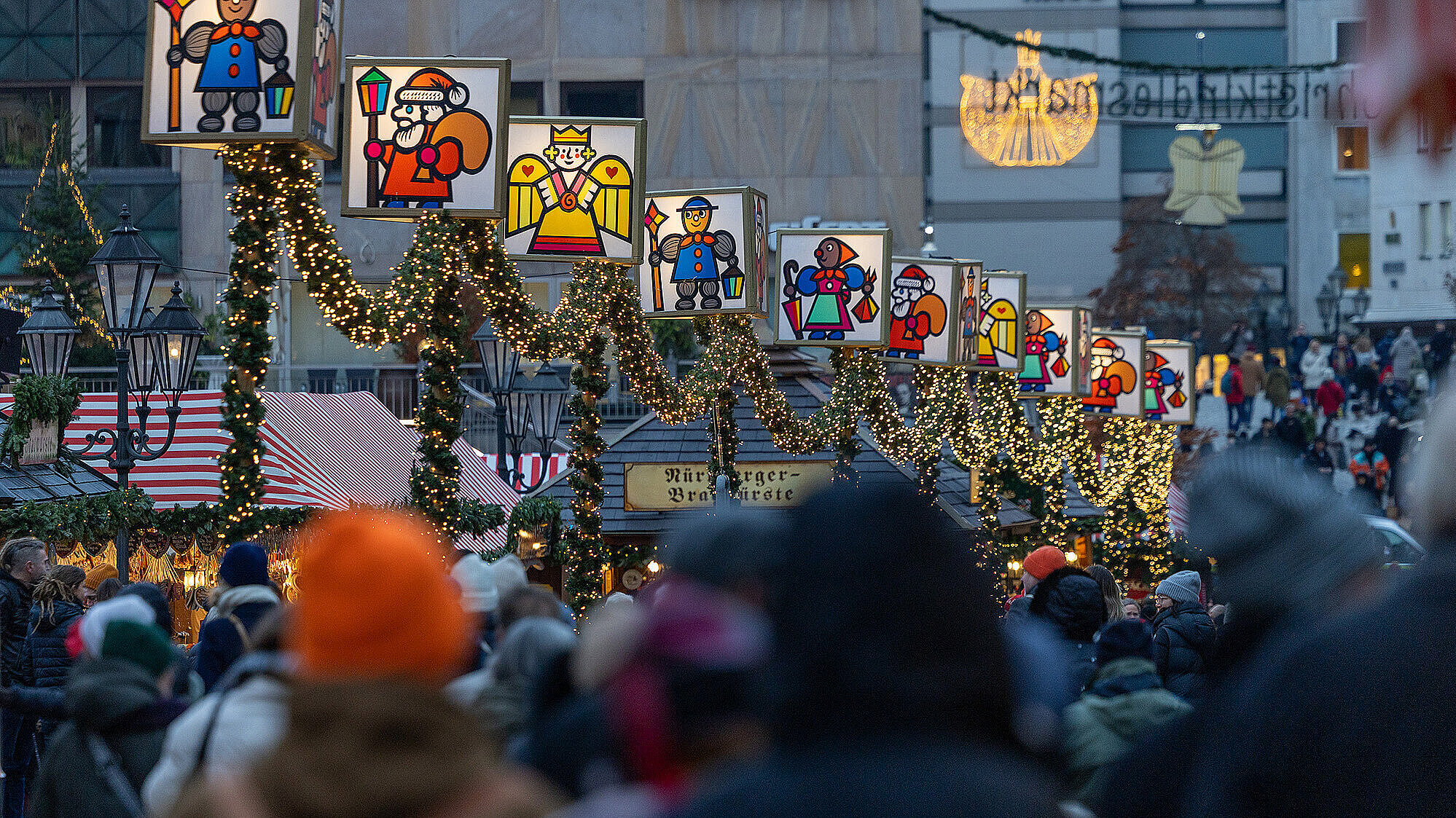 Menschenmenge auf dem Nürnberger Christkindlesmarkt bei Dämmerung, mit festlich geschmückten Ständen und beleuchteten Laternen in Form von traditionellen Weihnachtsfiguren, die an einer Girlande aus Tannenzweigen und Lichterketten hängen. Im Hintergrund ist ein Schild mit der Aufschrift „Nürnberger Bratwürste“ zu sehen.