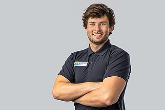 Max Kühnhauser vor hellem Hintergrund, trägt dunkles Poloshirt mit rechteckigem Aufnäher und hat die Arme verschränkt.
