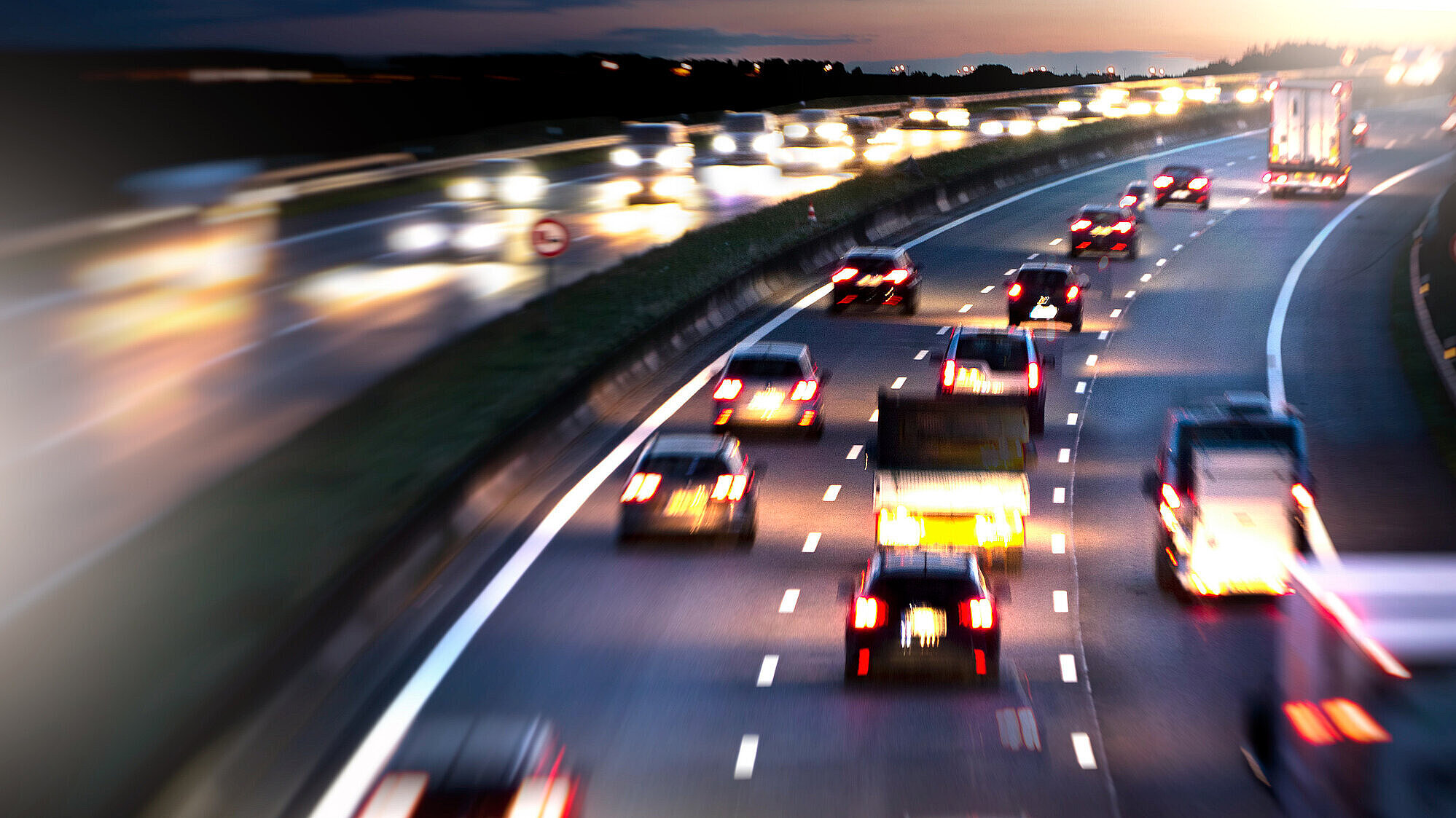 Auf dem Bild sind mehrere Autos auf der Autobahn zu sehen, bei Dämmerung. Alle Autos haben ihre Beleuchtung an.