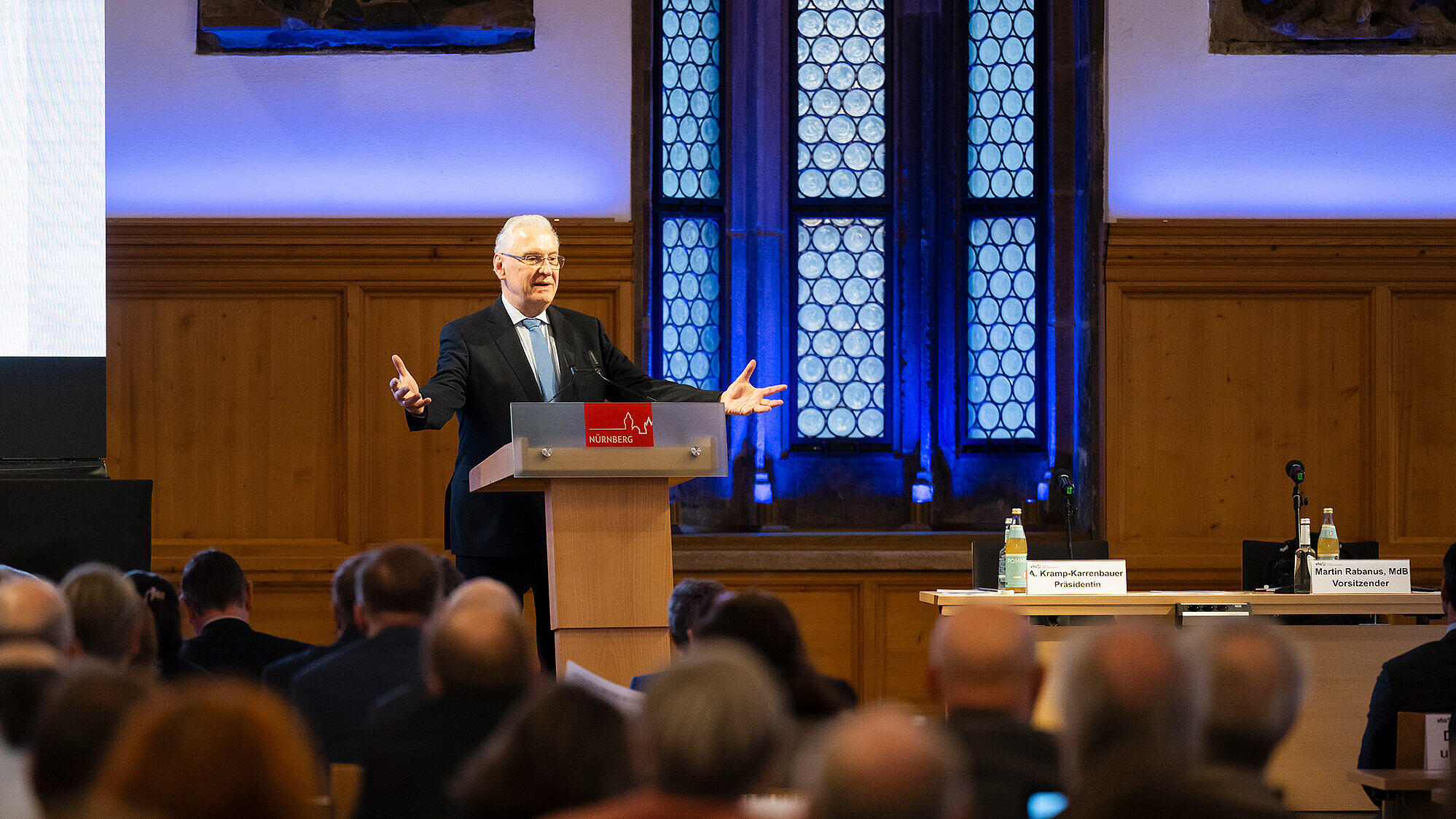Innenminister Joachim Herrmann dunklen Anzug steht an einem Rednerpult mit dem Schriftzug „Nürnberg“ und spricht vor einem Publikum in einem Saal mit hohen, bleiverglasten Fenstern und Holzvertäfelung. Rechts neben dem Rednerpult steht ein Tisch mit zwei Namensschildern, auf denen „A. Kramp-Karrenbauer Präsidentin“ und „Martin Rabanus, MdB Vorsitzender“ zu lesen sind. Das Publikum sitzt und hört aufmerksam zu.