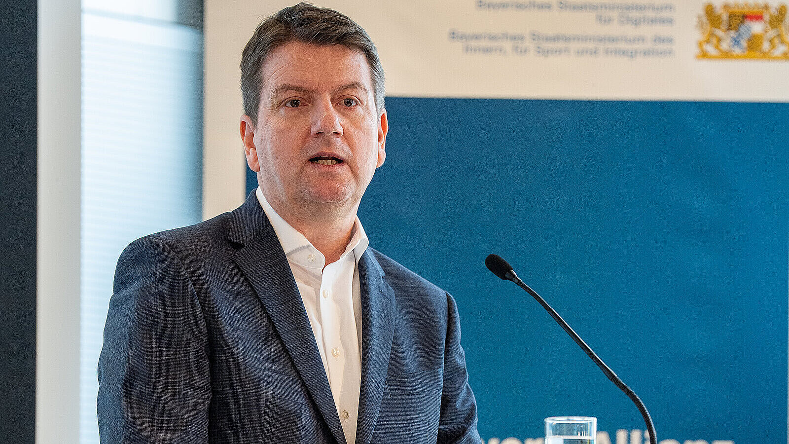 Innenstaatssekretär Sandro Kirchner redet an einem weißen Rednerpult mit StMI-Logo. Hinter ihm ein blaues Roll-Up mit Aufschrift "Bayern-Allianz gegen Desinformation"