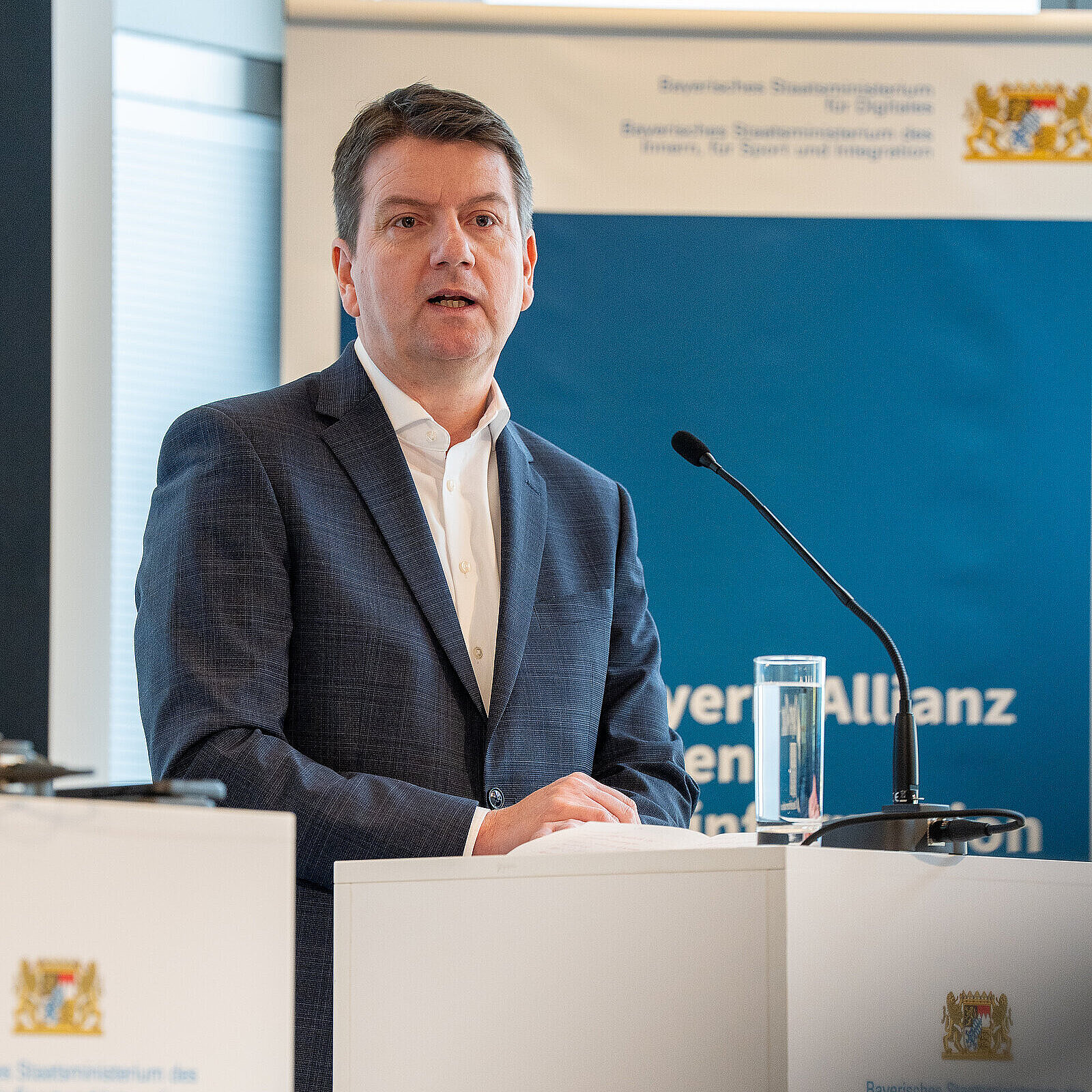 Innenstaatssekretär Sandro Kirchner redet an einem weißen Rednerpult mit StMI-Logo. Hinter ihm ein blaues Roll-Up mit Aufschrift "Bayern-Allianz gegen Desinformation"