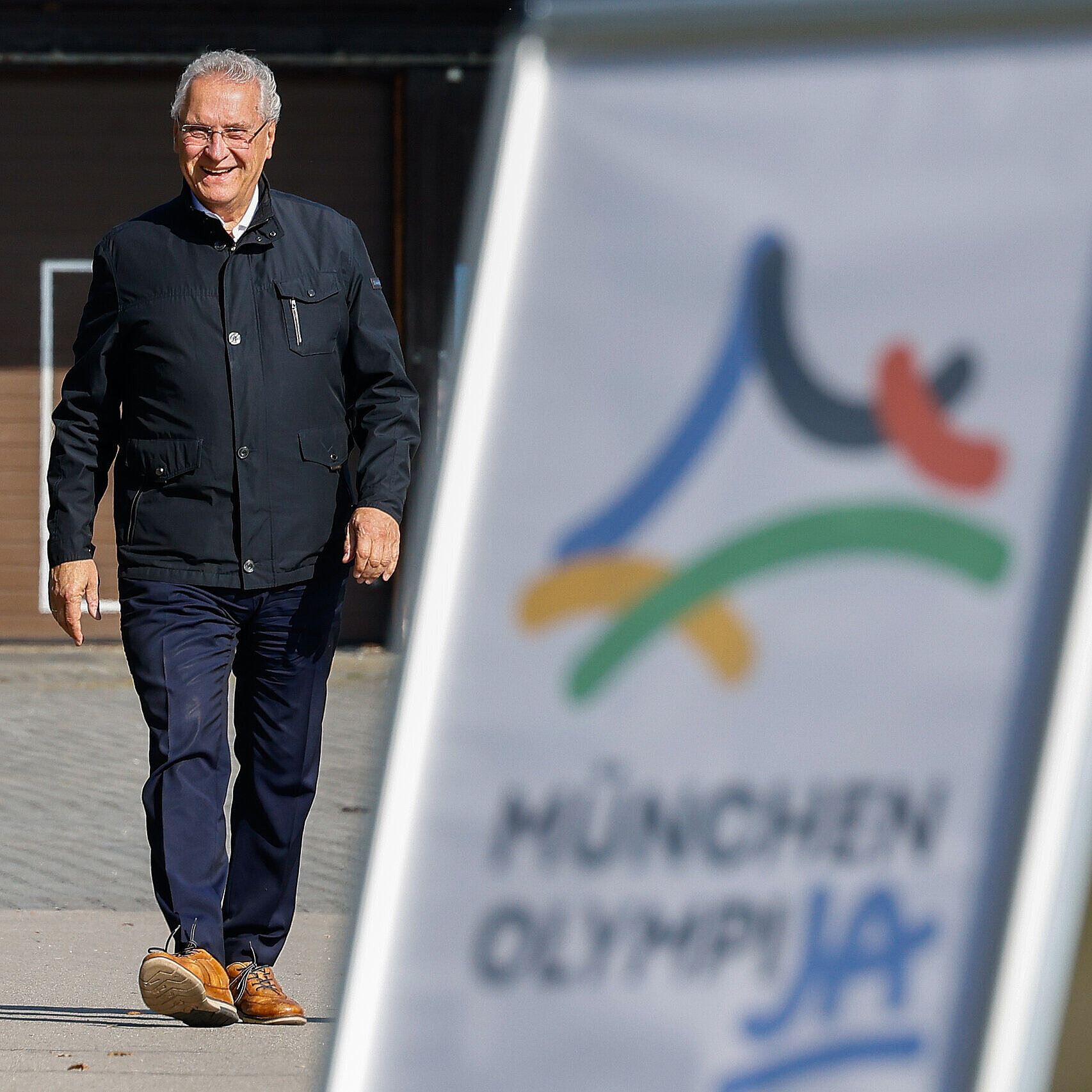 Staatsminister Joachim Herrmann gehend von vorne, im unscharfen Vordergrund Aufsteller mit OlympiJA-Logo