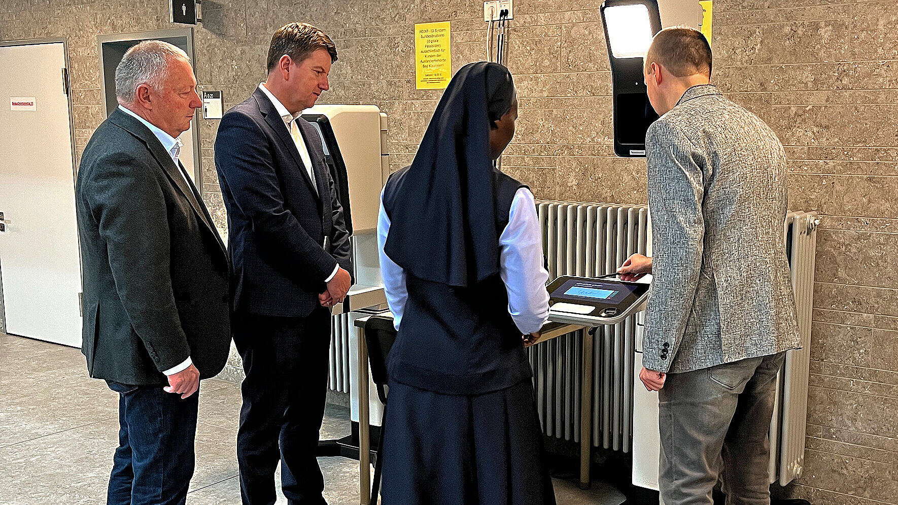 Auf dem Bild sind Innensstaatssekretär Sandro Kirchner, Landrat Thomas Bold, eine Kundin und ein Mitarbeiter des Landratsamtes zu sehen, die an einem elektrischen Unterschriften-Gerät stehen.