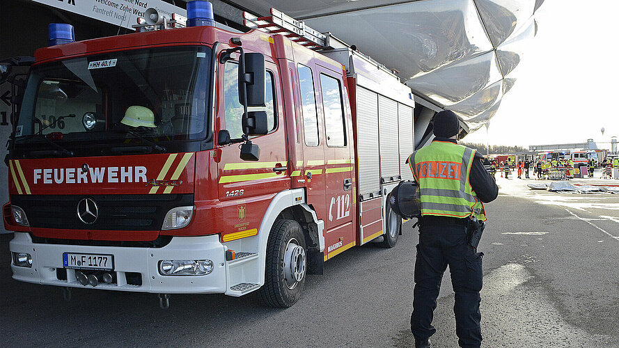 Ein Feuerwehrauto und ein Polizist stehen an einem Eingang der Allianz-Arena, der Polizist schaut auf einen Rettungseinsatz mit mehreren Personen und Rettungswagen im Hintergrund