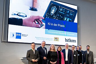 Gruppenfoto vor Präsentationsleinwand "KI in der Praxis" mit Innenminister Herrmann und weitere Akteure der Veranstaltung