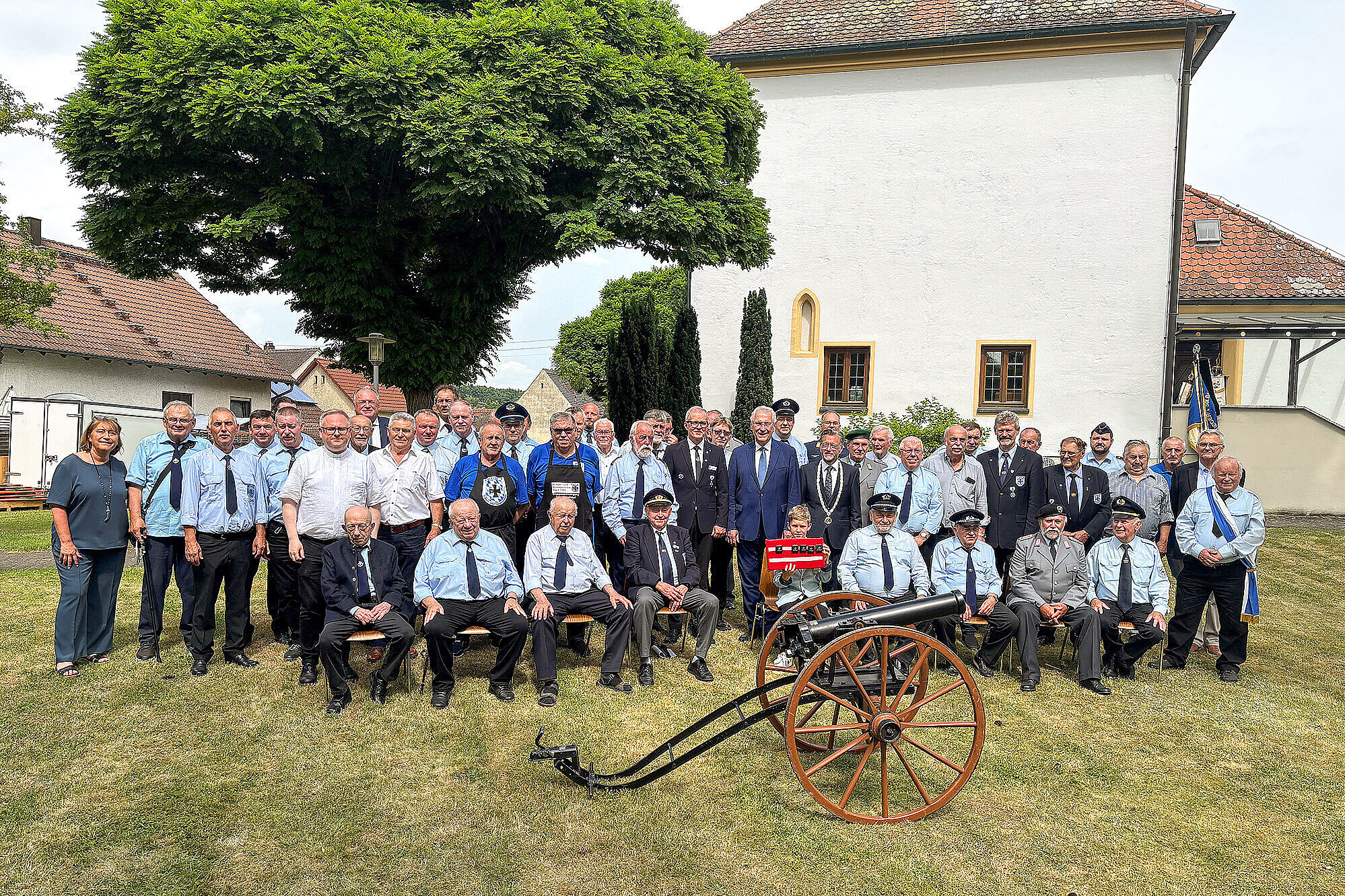 Gruppenfoto von rund 50 Personen bei einer Gedenk- oder Veteranenveranstaltung im Freien. Die meisten Männer tragen Uniformen oder festliche Kleidung, einige mit Schärpen oder Abzeichen. Vorne sitzen ältere Herren auf Bänken, davor steht eine historische Kanone. Im Hintergrund ein Baum, Rasenfläche und ein helles Gebäude mit roten Ziegeldächern. Ein Kind hält eine kleine Deutschlandfahne mit „150 Jahre“ hoch. Feierliche Stimmung bei sommerlichem Wetter.
