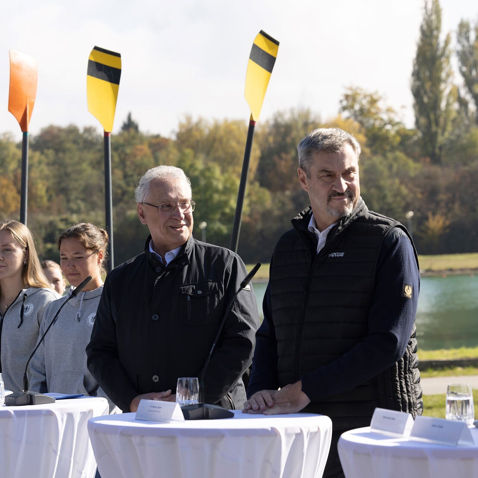 Auf dem Bild sind Sportminister Joachim Herrmann und Ministerpräsident Dr. Markus Söder zu sehen während der PK hinter Stehtischen. Dahinter sind drei junge Ruderinnen mit Rudern zu sehen.