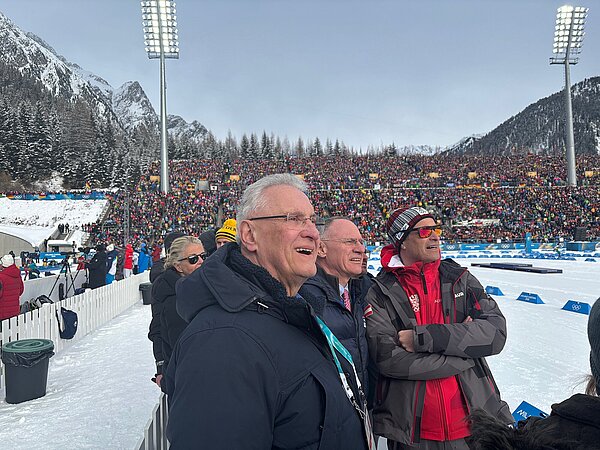 Zuschauer in Winterjacken lehnen an einer Absperrung und beobachten einen Skilanglauf‑Wettkampf auf verschneiter Strecke; Tribüne mit Zuschauern und Olympia‑Logos im Hintergrund