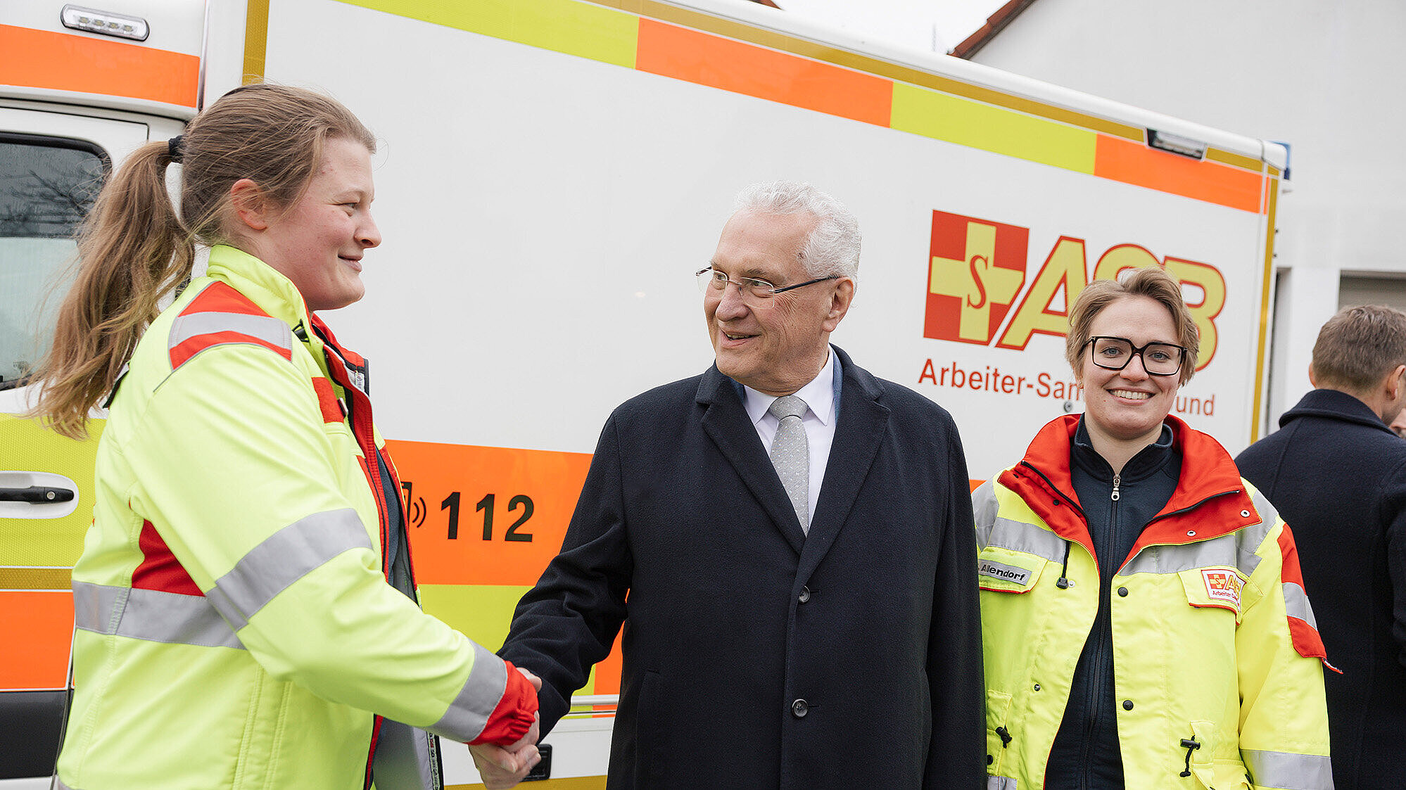 Innenminister Joachim Herrmann in einem schwarzen Mantel und mit grauen Haaren schüttelt die Hand einer Frau in einer gelb-roten Rettungsdienstjacke. Neben ihnen steht eine weitere Person in einer ähnlichen Rettungsdienstjacke. Im Hintergrund ist ein Rettungswagen mit dem Schriftzug „ASB Arbeiter-Samariter-Bund“ zu sehen.