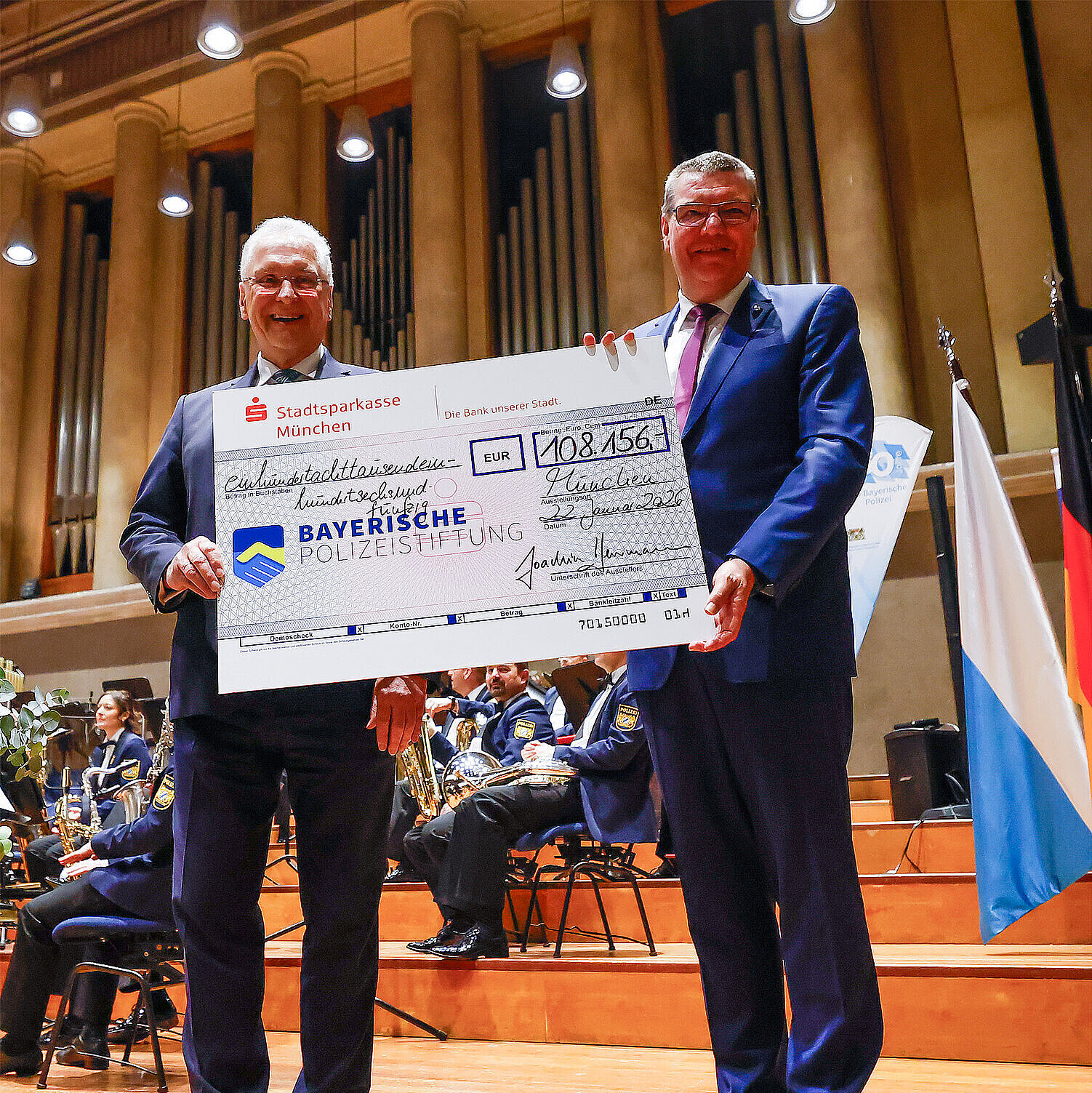 Zwei Männer in Anzügen stehen auf einer Bühne in einem Konzertsaal und halten gemeinsam einen großen symbolischen Spendenscheck der Stadtsparkasse München über 108.156 Euro zugunsten der Bayerischen Polizeistiftung. Im Hintergrund sind Musiker in Uniformen mit Blasinstrumenten zu sehen, sowie eine große Orgel und eine Bayerische Flagge.