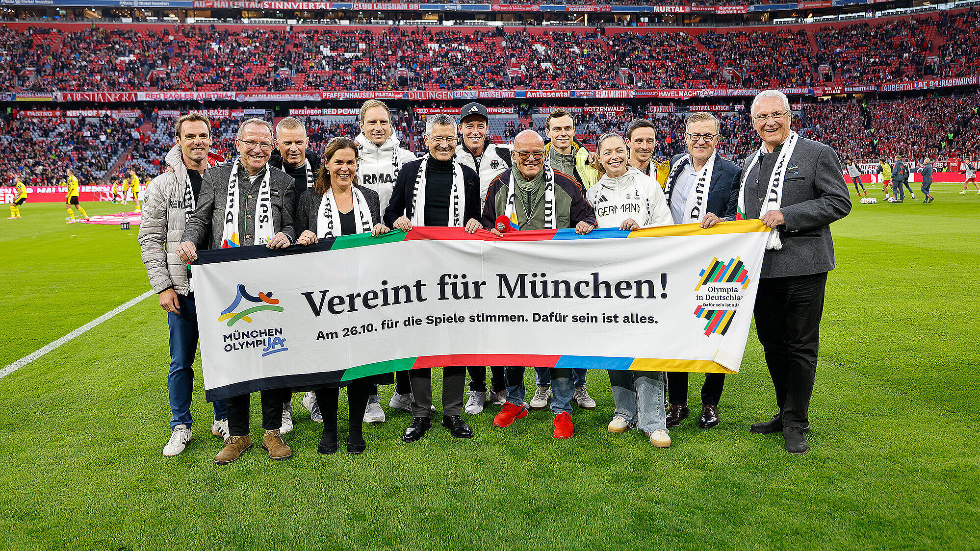 Das Bild zeigt eine Gruppe von Menschen auf einem Fußballfeld, die gemeinsam ein Banner halten. Im Hintergrund ist ein vollbesetztes Stadion mit Zuschauern zu sehen. Die Personen tragen Schals und Kleidung, die auf eine Unterstützung für München hinweisen, und das Banner enthält den Text "Verein für München! Am 26.10. für die Spiele stimmen. Dafür sein ist alles."