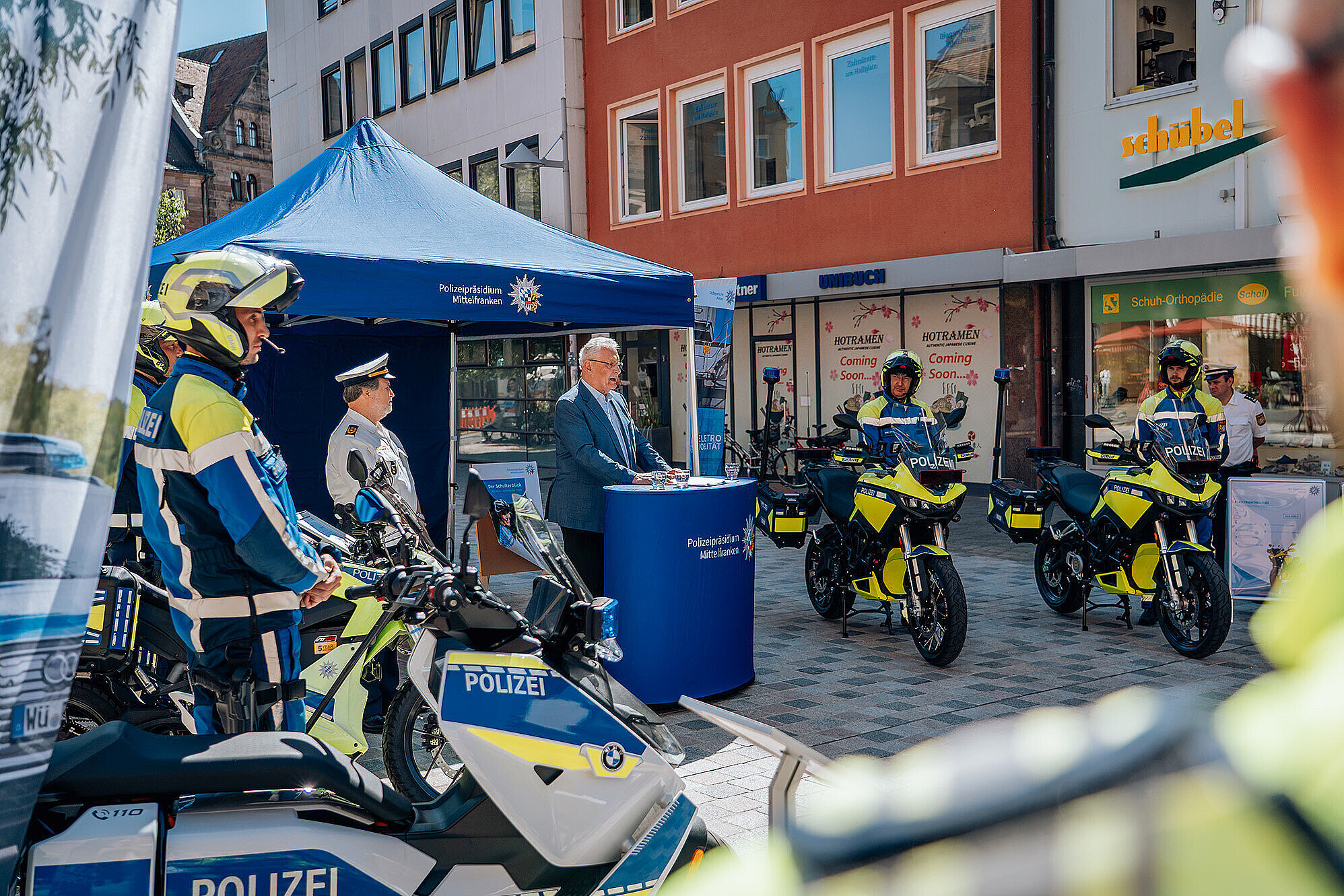 Ein Polizeistand mit mehreren Einsatzfahrzeugen und Polizisten in Uniform, Innenminister Joachim Herrmann am Rednerpult vor einem blauen Zelt,auf einer Stadtstraße