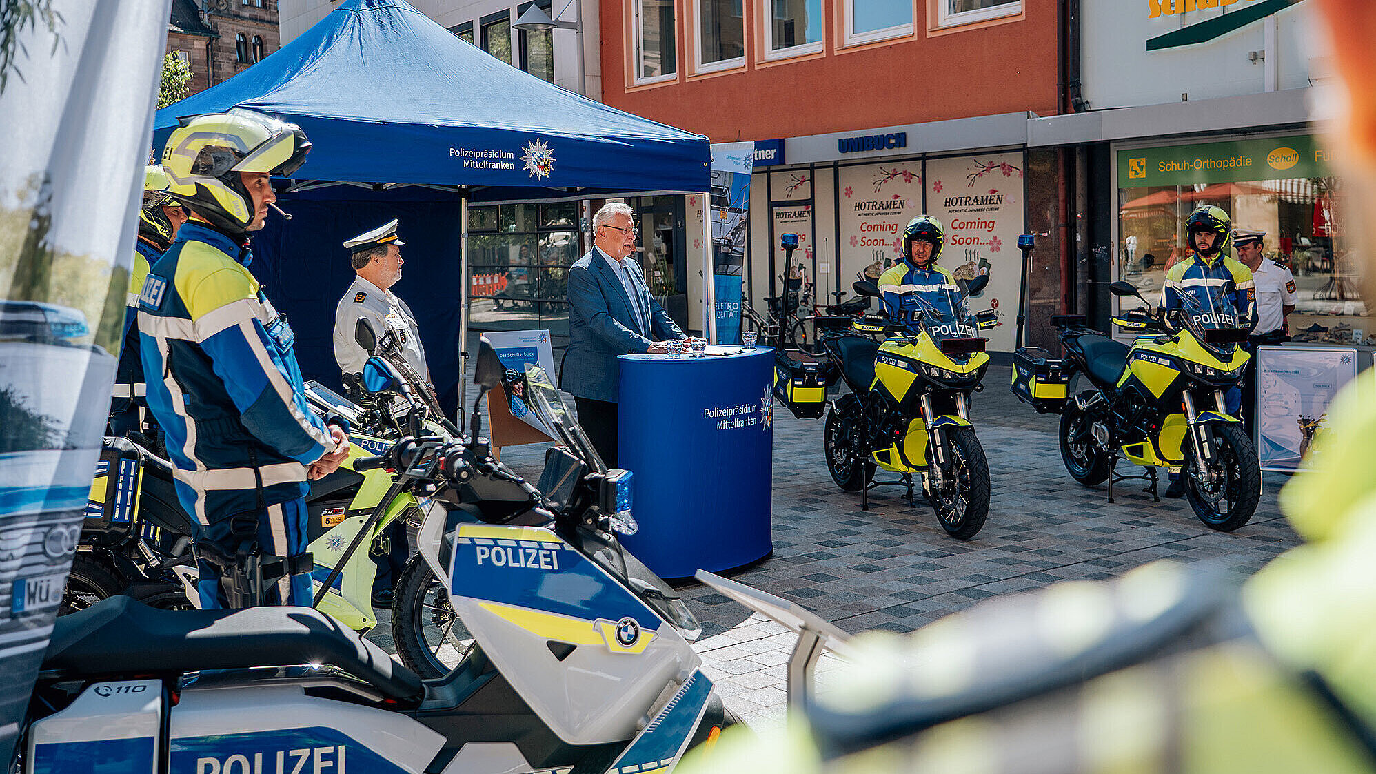 Ein Polizeistand mit mehreren Einsatzfahrzeugen und Polizisten in Uniform, Innenminister Joachim Herrmann am Rednerpult vor einem blauen Zelt,auf einer Stadtstraße