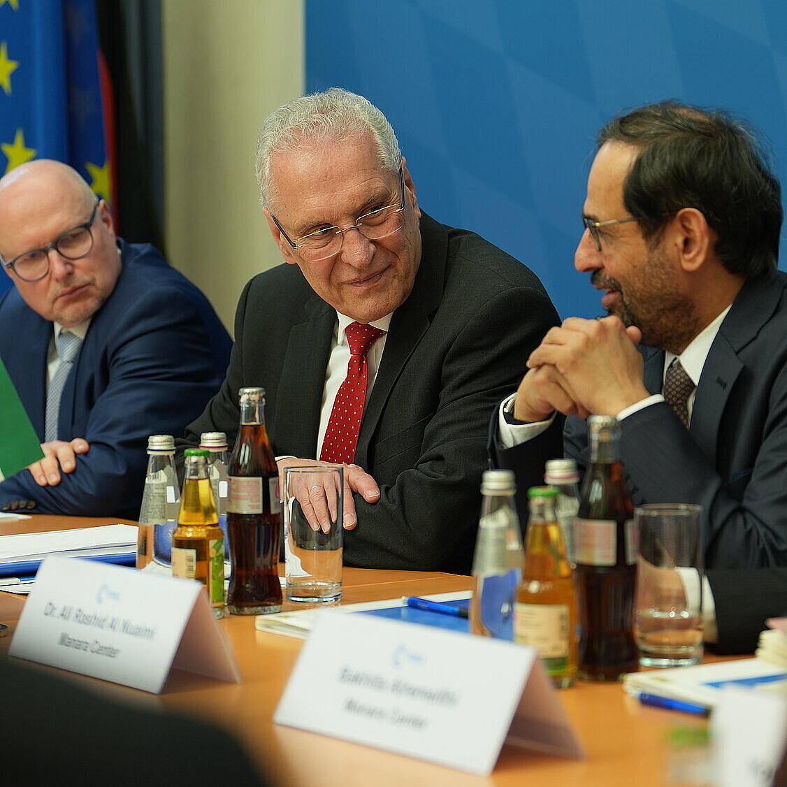 Mehrere Personen in Anzug sitzen an einem Konferenztisch in einem Besprechungsraum. Auf dem Tisch stehen zahlreiche Flaschen mit Getränken, Gläser, Schreibutensilien und Namensschilder. Im Hintergrund sind eine blaue Wand mit weiß-blauem Rautenmuster sowie europäische und deutsche Flaggen zu sehen.