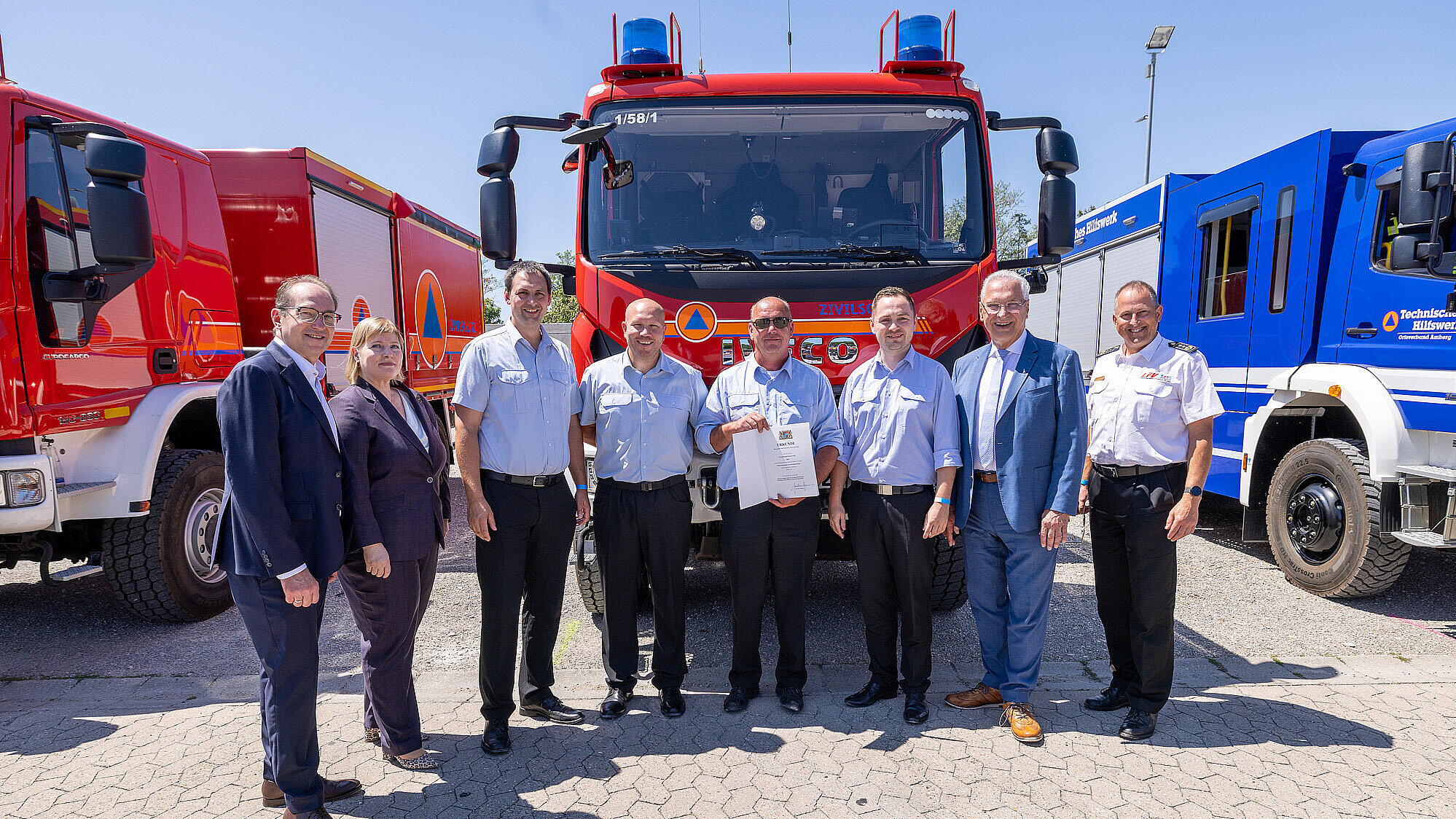 Neun Personen — größtenteils Einsatzkräfte in hellblauen Hemden, zwei in Anzügen — stehen in einer Reihe vor einem roten Einsatz-Lkw (neben weiteren roten und blauen Fahrzeugen). Eine Person hält eine Urkunde; sonniger Außenbereich.