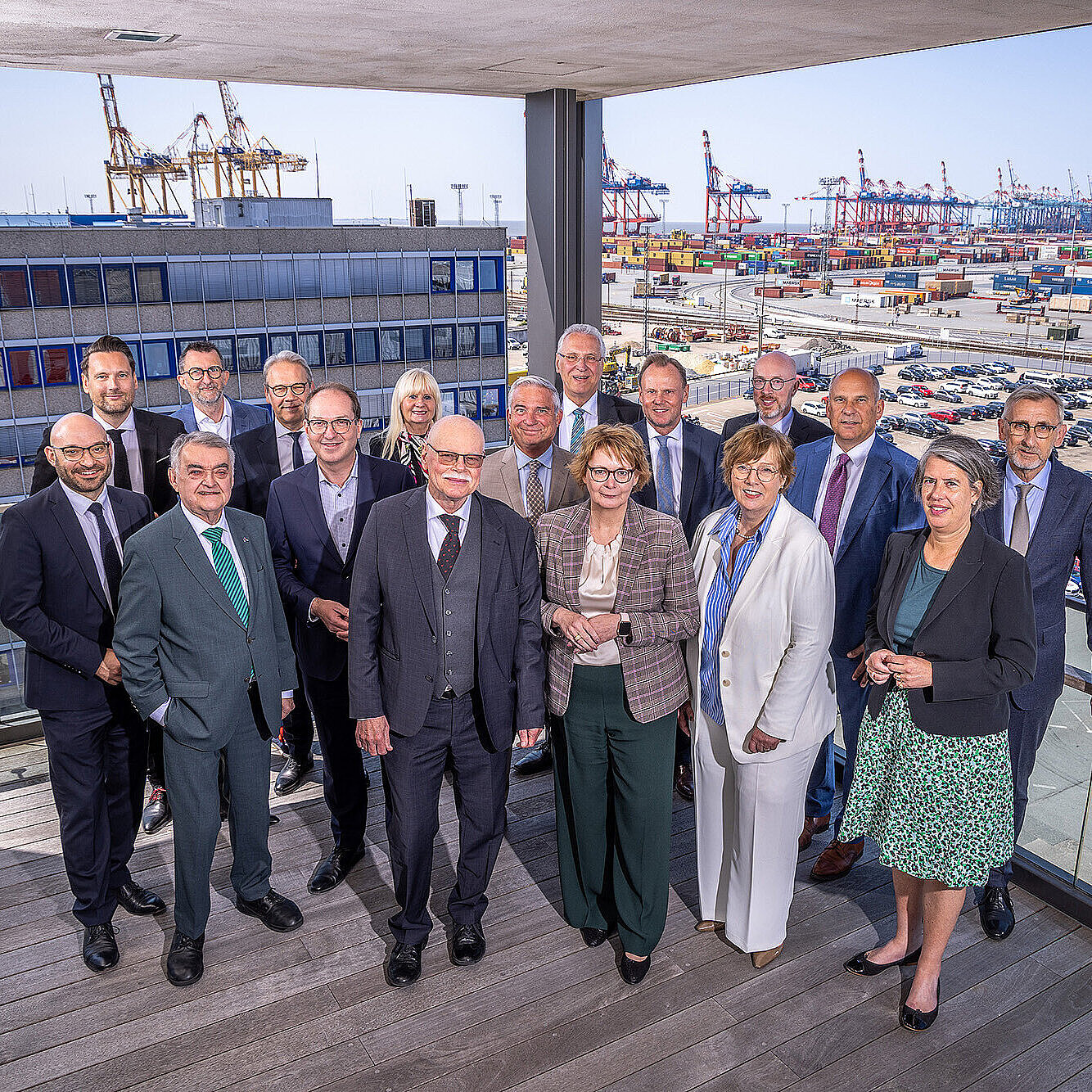 Gruppenfoto der Teilnehmer der IMK Personen in Businesskleidung auf einem Balkon mit Blick auf einen Containerhafen. Im Hintergrund sind zahlreiche Container, Kräne und geparkte Autos zu sehen. Die Personen stehen eng beieinander und lächeln in die Kamera. 