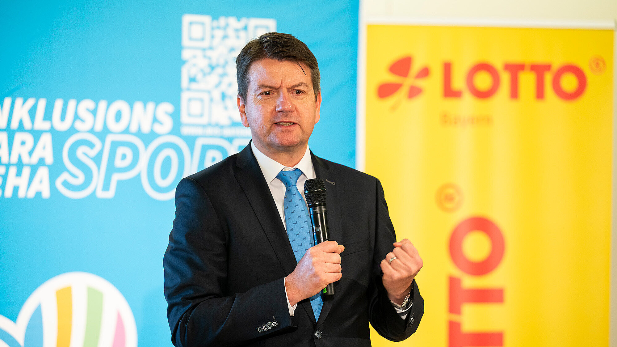 Staatssekretär Sandro Kirchner in einem dunklen Anzug mit weißem Hemd und blauer Krawatte hält ein Mikrofon in der Hand und spricht bei einer Veranstaltung. Im Hintergrund sind Logos und Banner mit den Worten "LOTTO" und "SPORT" sowie ein QR-Code zu sehen.